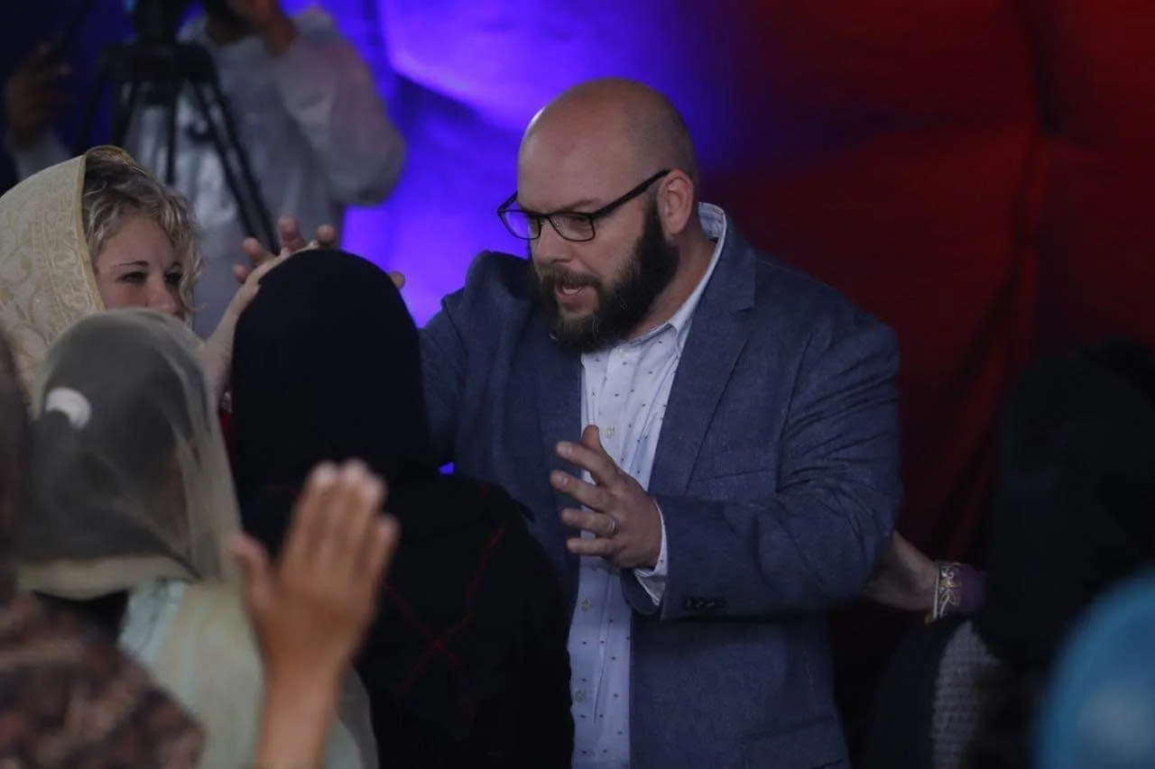 A man with glasses and a beard, dressed in a blue blazer and white shirt, is praying or speaking with several women, some wearing headscarves, in a dimly lit room with purple and red lighting.