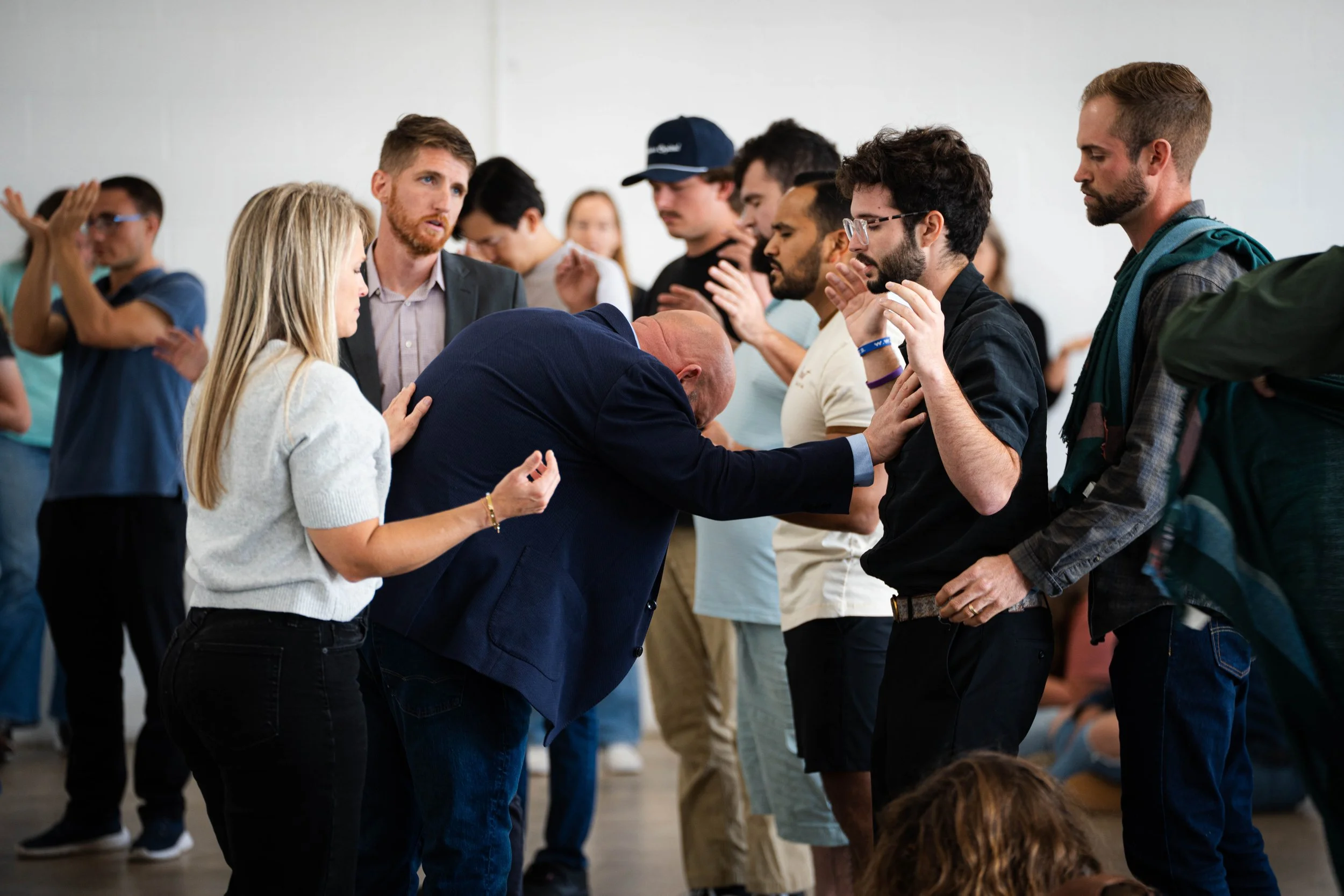 A group of people standing in a circle, praying or meditating with their eyes closed, some with their hands raised or clasped, in a peaceful indoor setting.