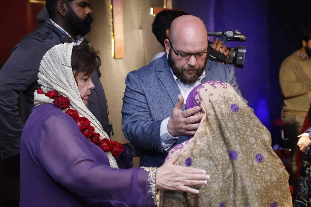 A woman dressed in purple with a red flower necklace and a scarf, holding a hand on a woman in a patterned shawl. A man wearing a blue suit, glasses, and a beard is gently holding and blessing the woman in shawl during a ceremony.