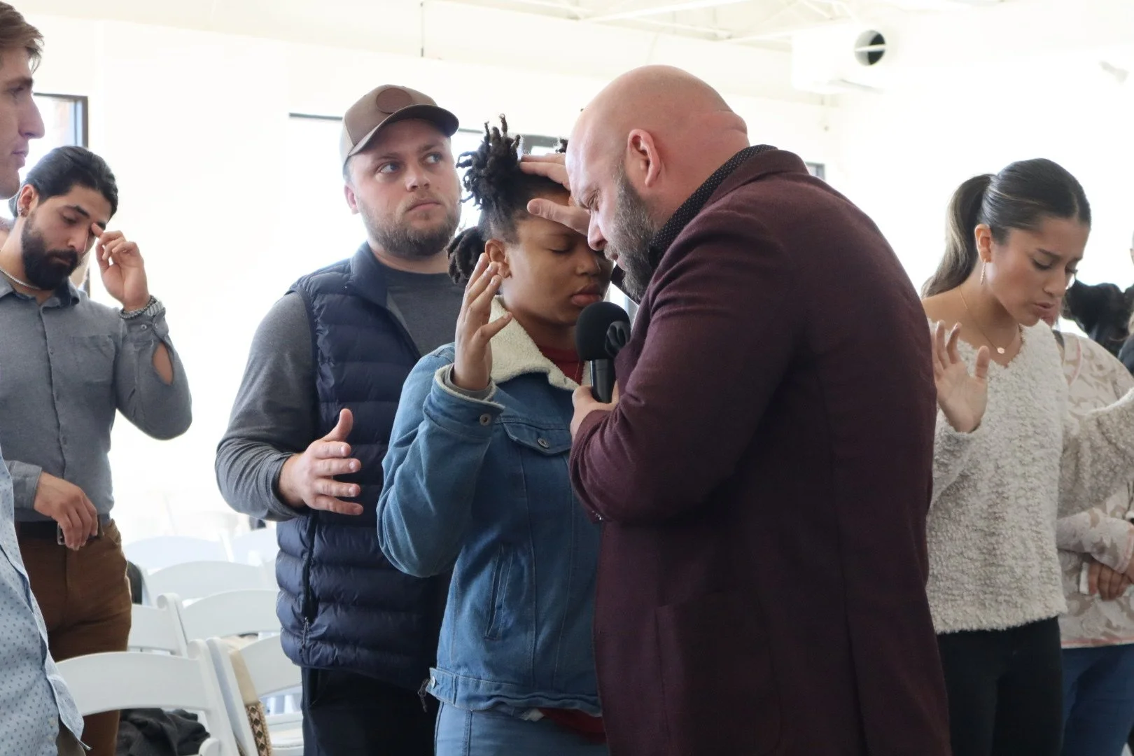 A group of people praying with their hands raised, including a woman with dreadlocks and a man with a beard holding a microphone, in an indoor setting.