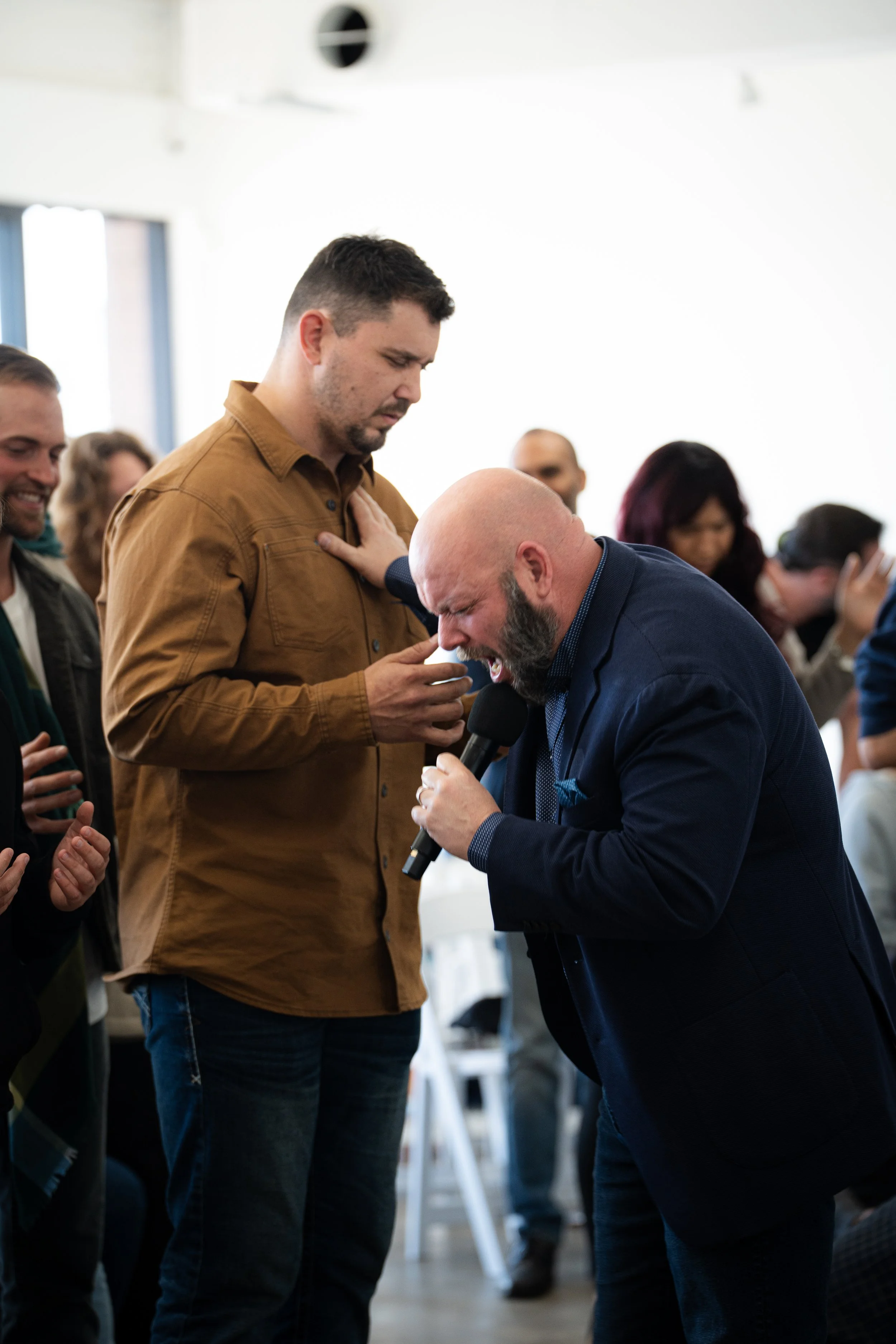 A man in a brown shirt appears to be praying with his hand on his chest while a bald man in a dark suit prays with a microphone in hand at a religious gathering.