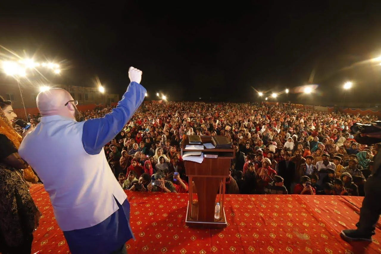 A man at a podium addressing a large crowd of people gathered at an outdoor event at night. The audience is dense and diverse, with many women wearing headscarves. There are bright lights illuminating the scene.