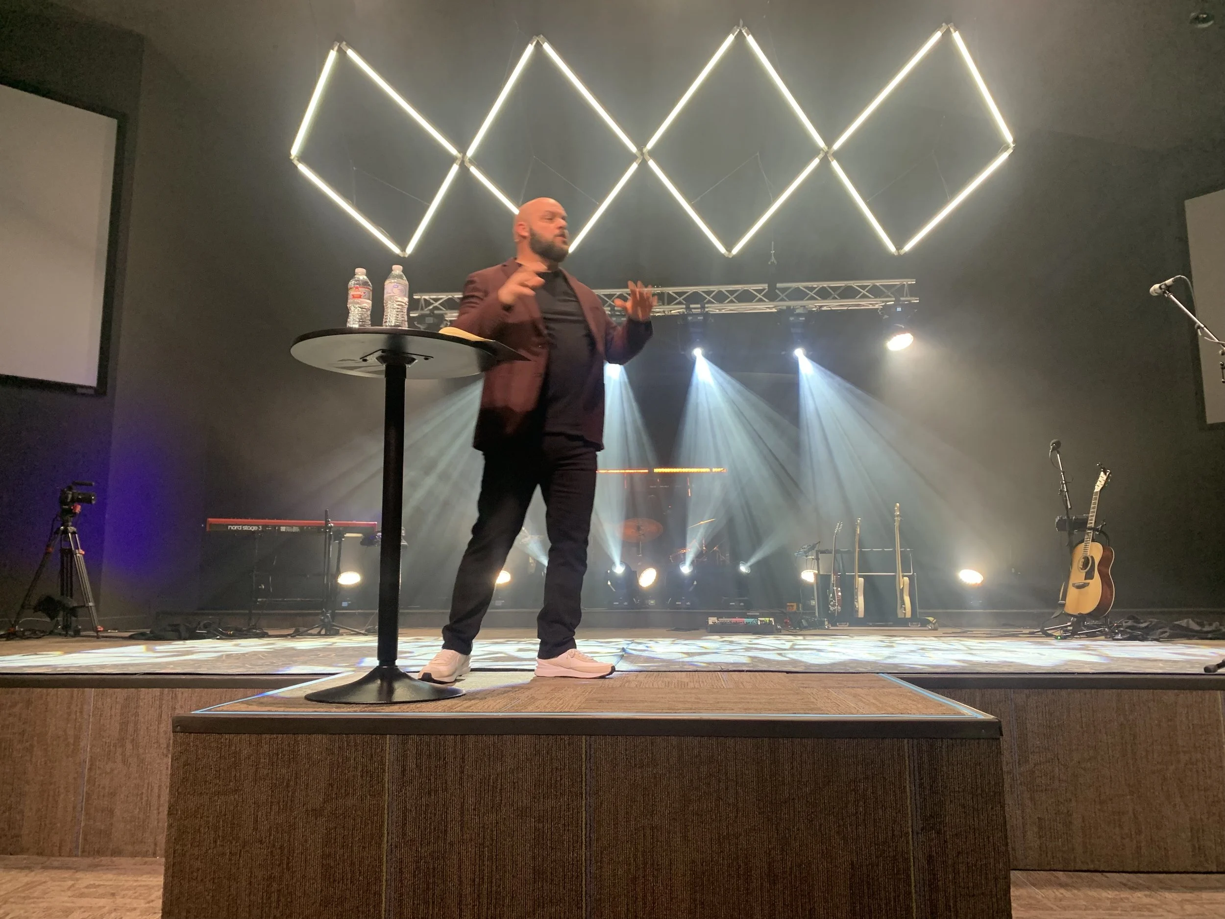 A man stands on a stage with musical instruments behind him, illuminated by stage lights, with a geometric light fixture overhead, and two water bottles on a small round table.
