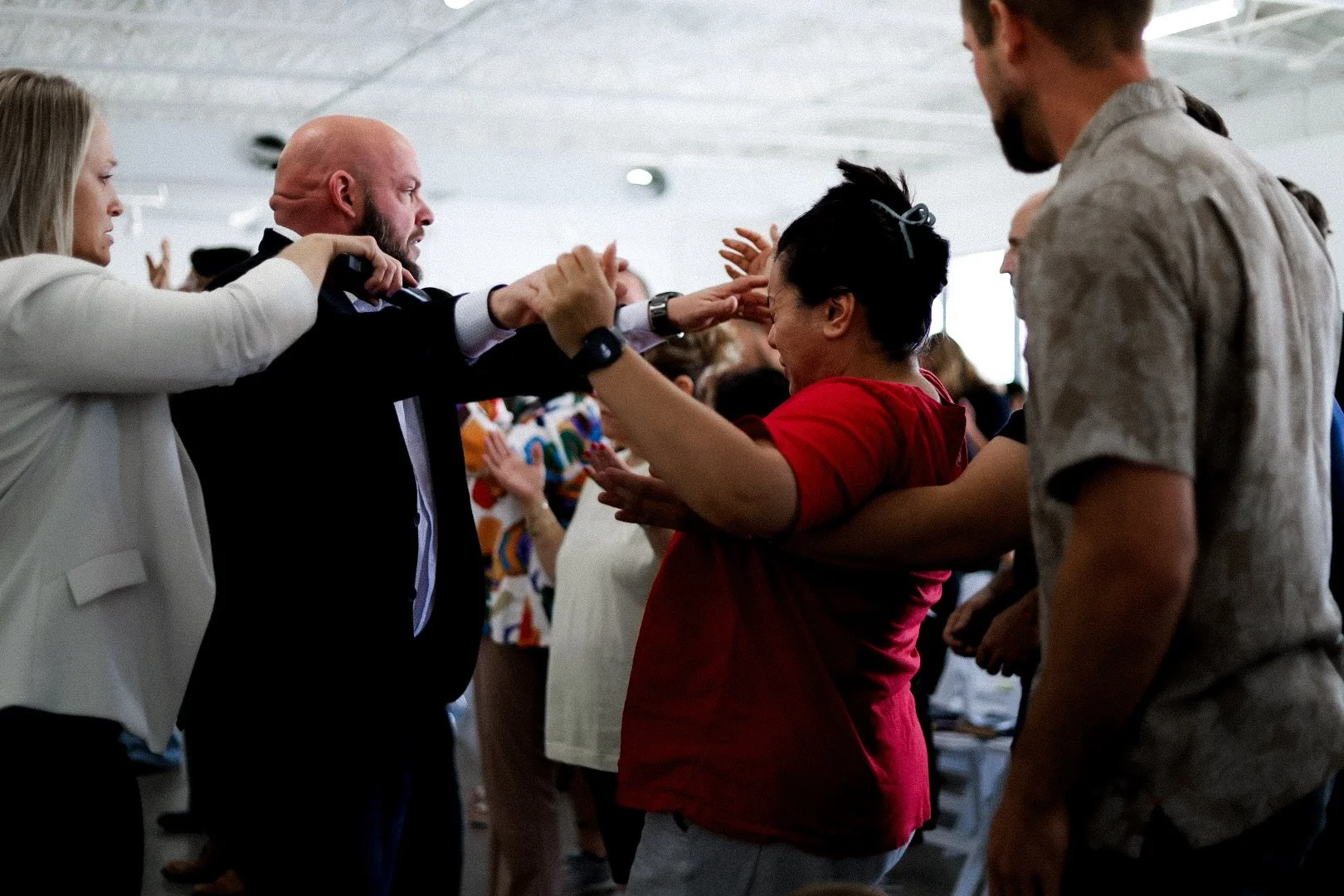 People participating in a faith healing event, with a man in a dark suit and a white shirt praying over a woman in a red shirt, others around them with hands raised.