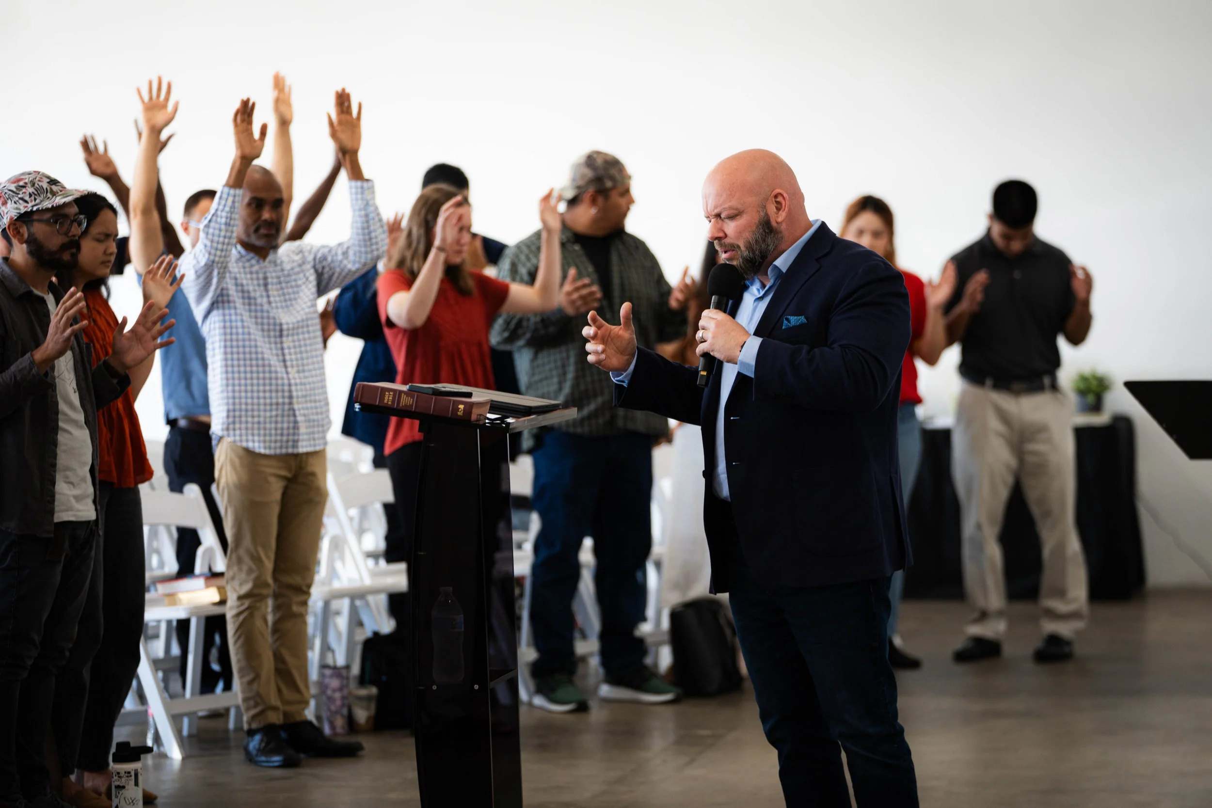 A man in a dark suit leading prayer in a church with people standing and raising their hands in prayer.