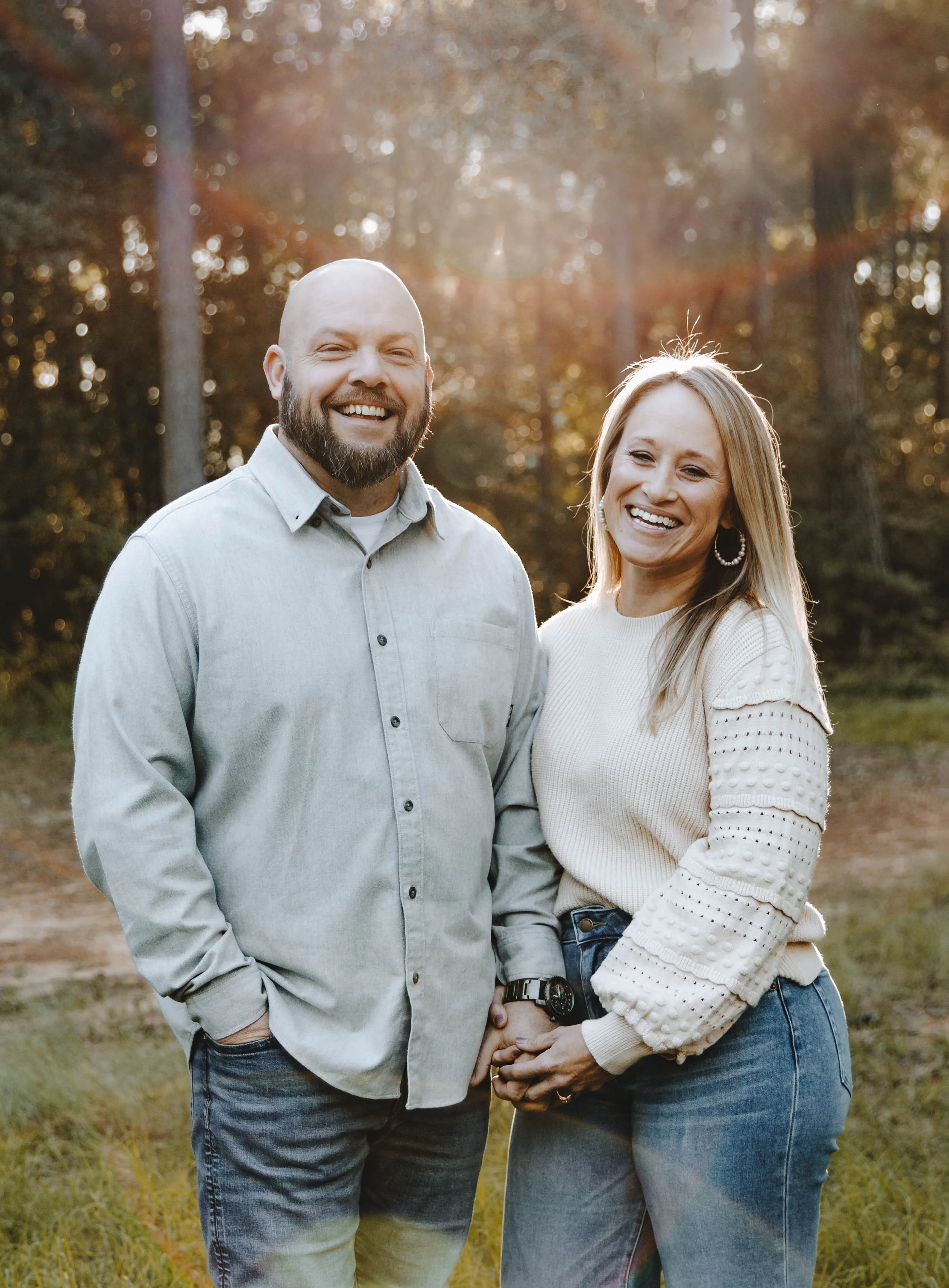 A smiling couple holding hands outdoors in a wooded area with sunlight filtering through the trees.