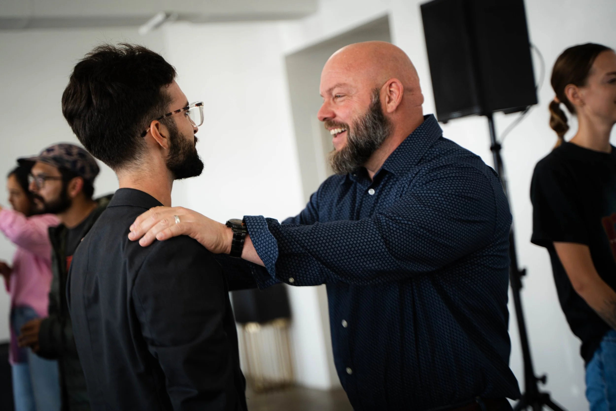 Two men are smiling and shaking hands at an indoor event. One has glasses and dark hair, while the other is bald with a beard. They appear to be greeting or congratulating each other.