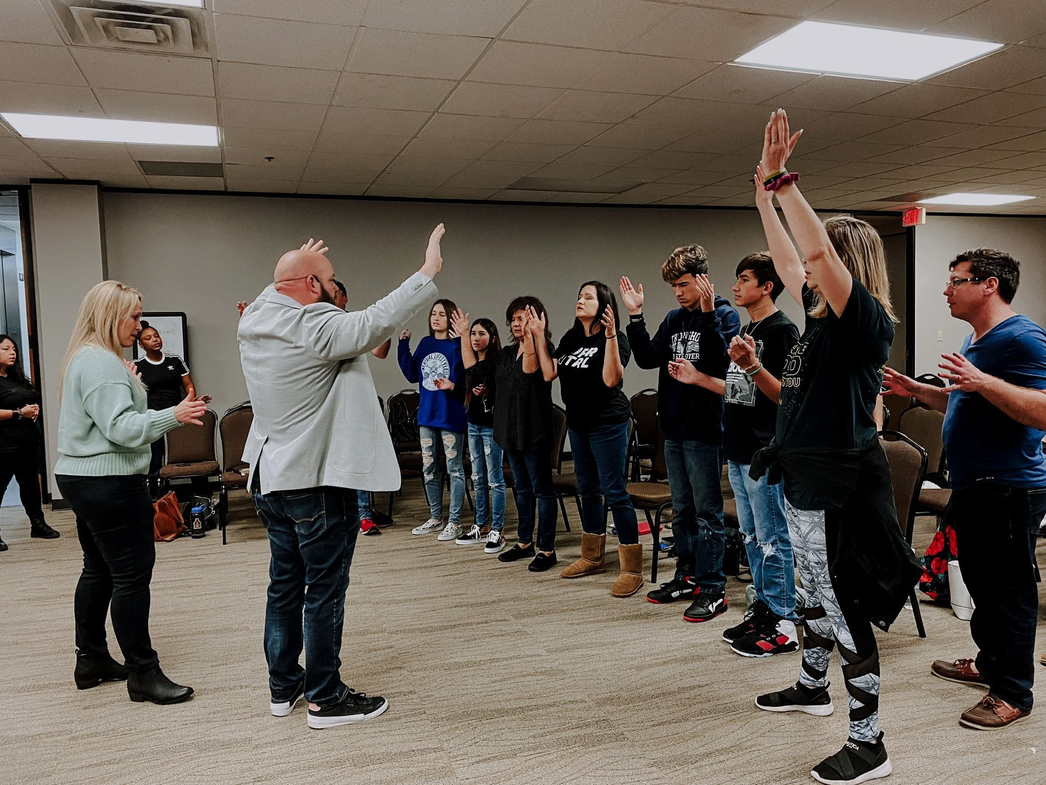 Group of people standing in a circle, participating in a discussion or activity, with some raising their hands in a room with beige walls and ceiling lights.