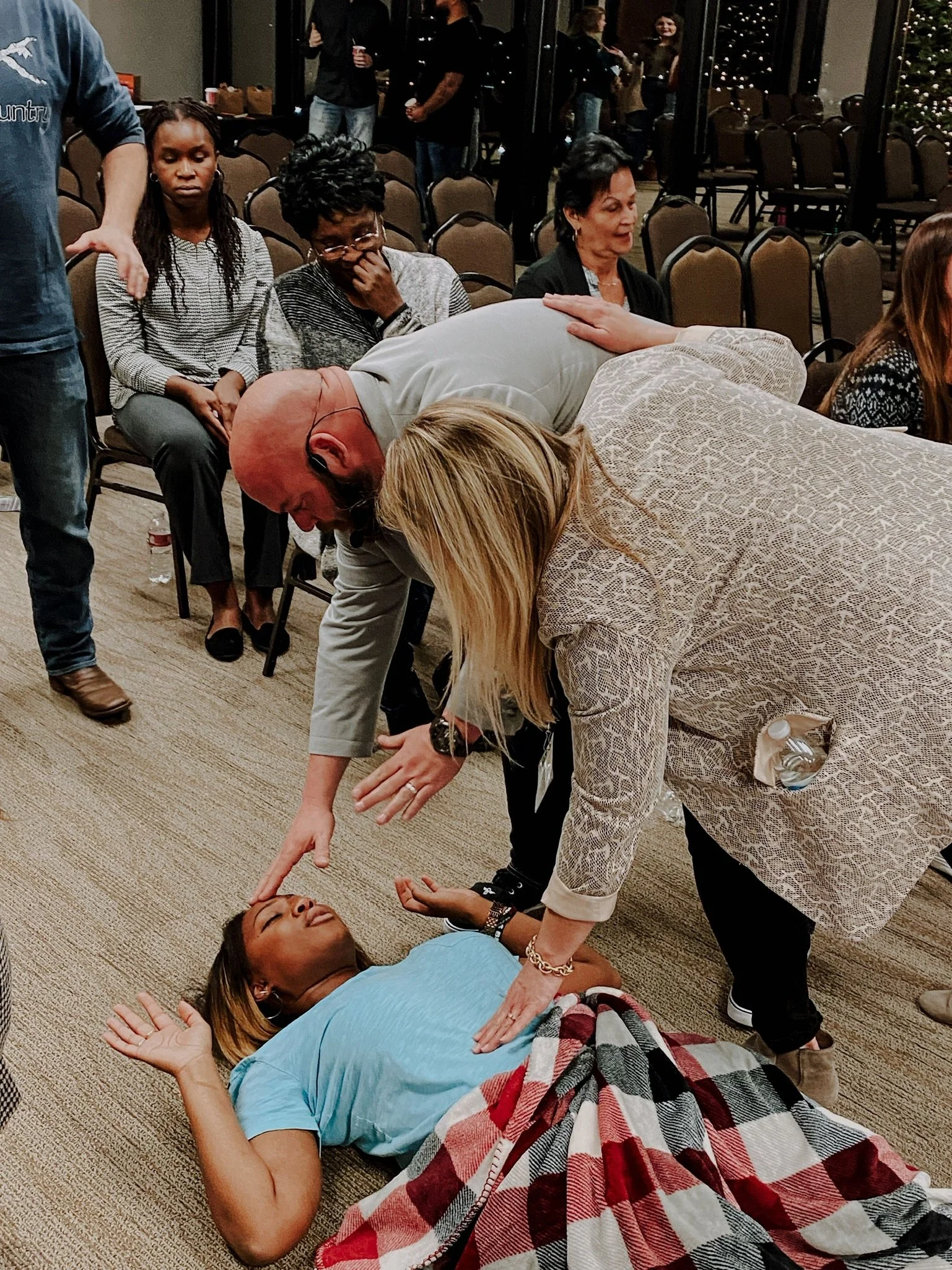 A woman lying on the floor in a blue shirt appears to be receiving CPR or first aid from a woman in a patterned blazer. Several people are seated nearby, with some observing and others preoccupied.
