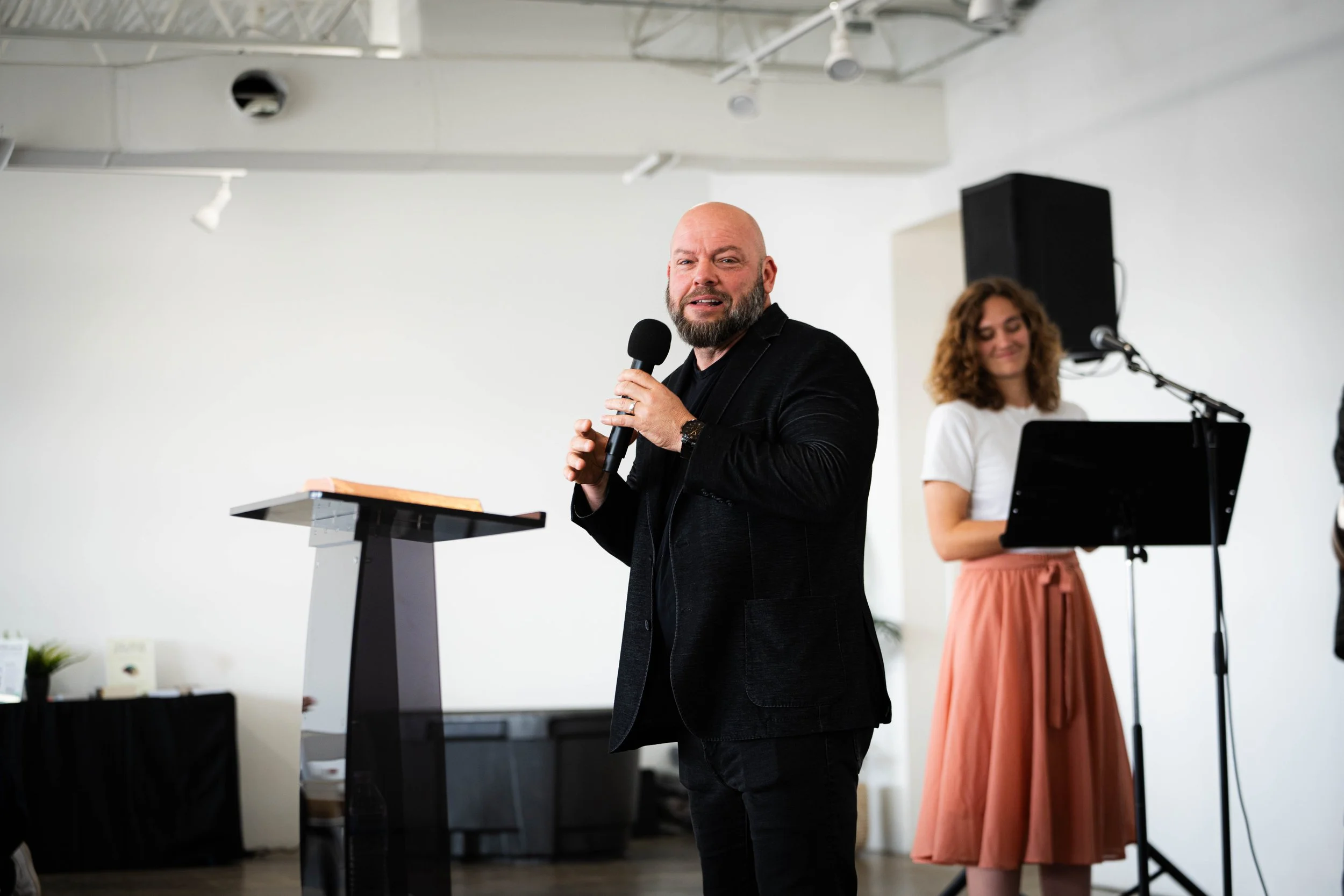 A man speaking into a microphone at a podium during an event, with a woman standing behind him near a music stand and speaker in a minimalistic room.