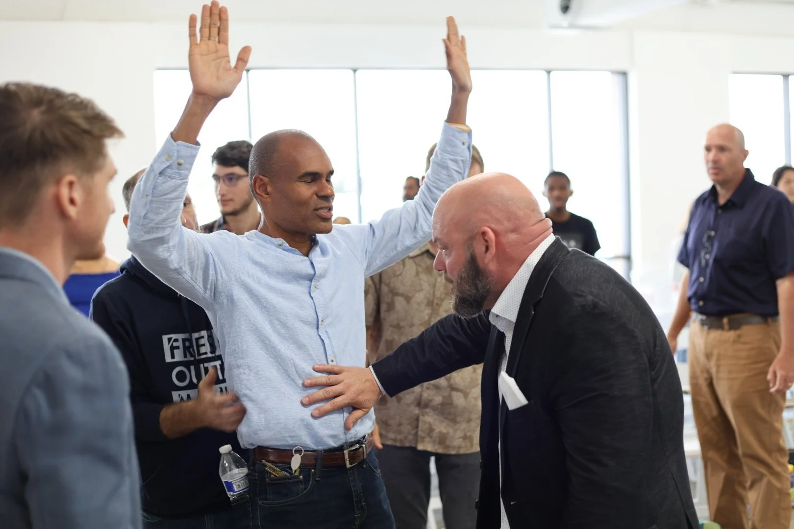 A diverse group of people participating in a spiritual or religious gathering, with two men at the center engaged in prayer or ritual, one with raised hands and the other with a hand on his chest.