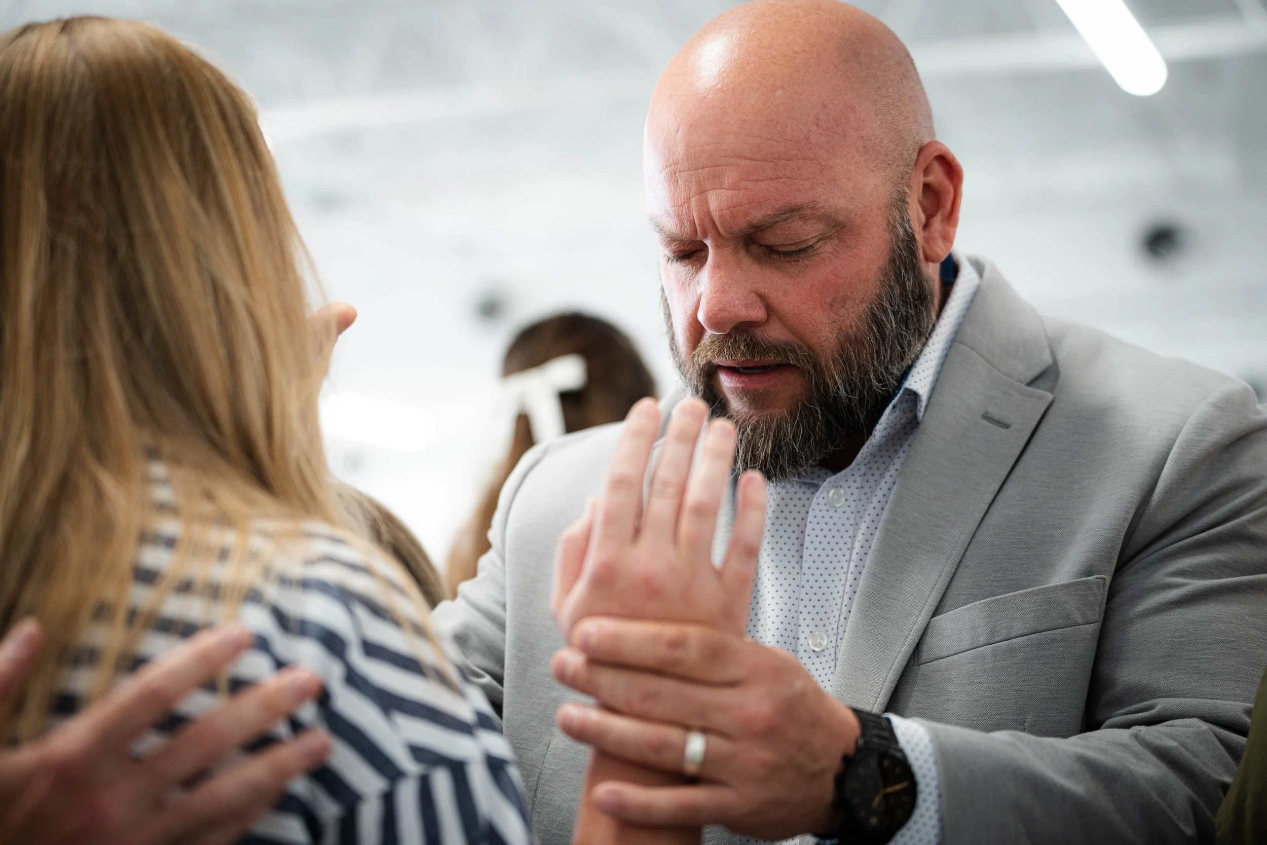 A bearded man in a light gray suit prays with eyes closed while touching a woman's hand.