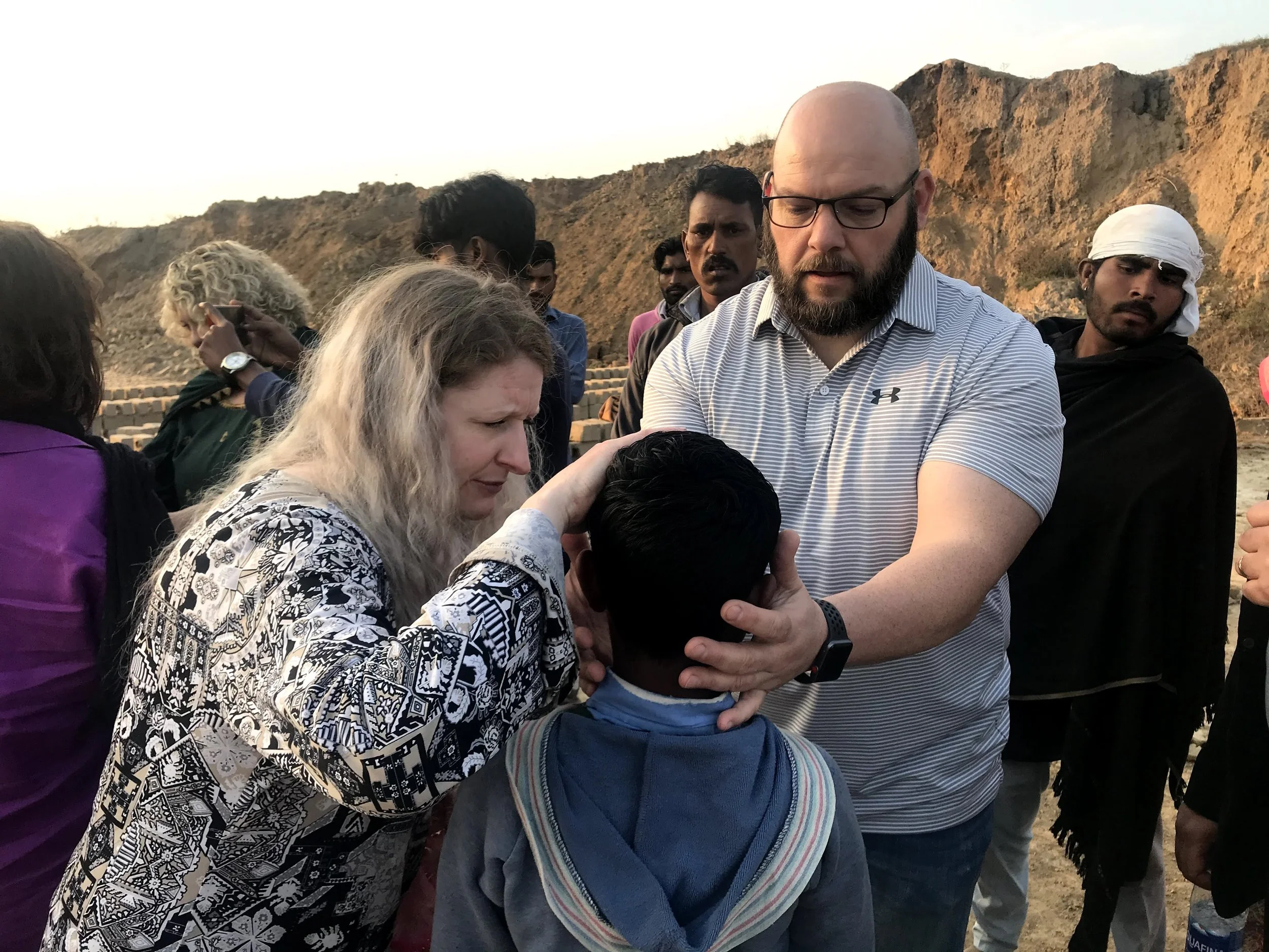 A group of people gather outdoors at the base of a rocky hill in a desert-like setting during sunset, with some individuals appearing to pray or engage in a spiritual activity.