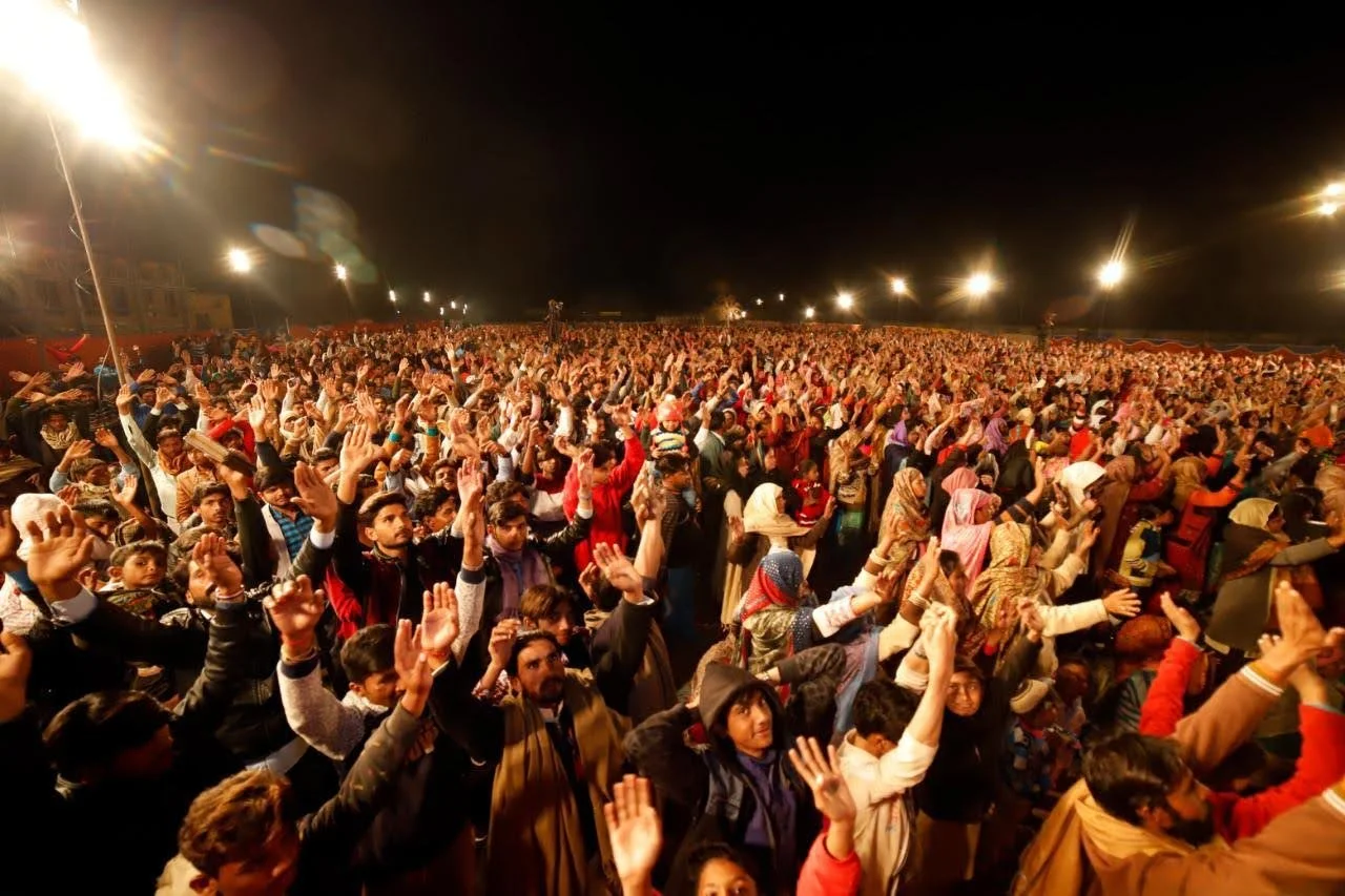 A large crowd of people gathered outdoors at night, raising their hands in the air, illuminated by bright streetlights.