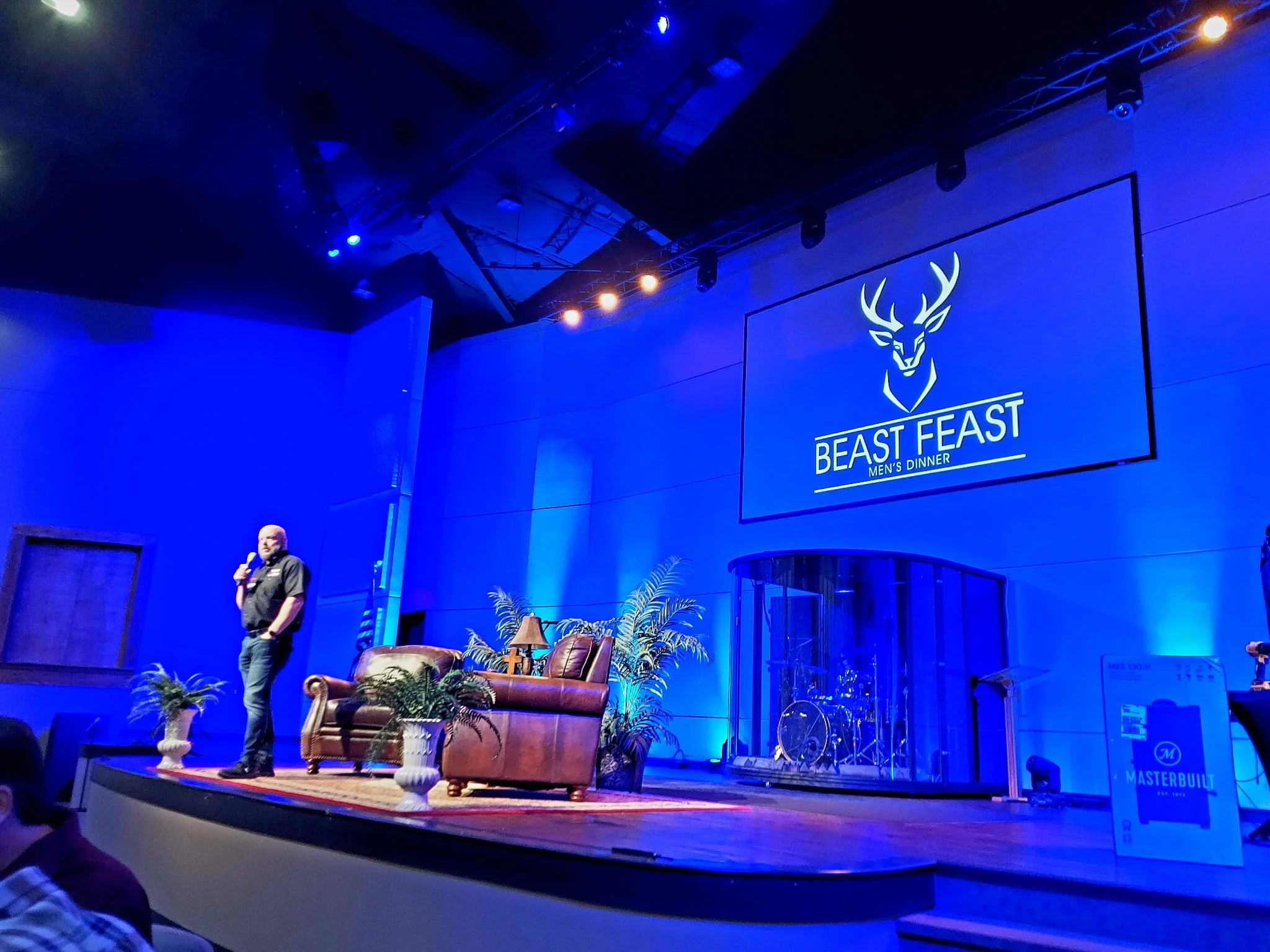A man standing on stage holding a microphone at a men's dinner event called Beast Feast. The stage includes a brown leather couch, potted plants, a small table with a lamp, and a drum set enclosed in glass. A large screen behind displays a logo with 