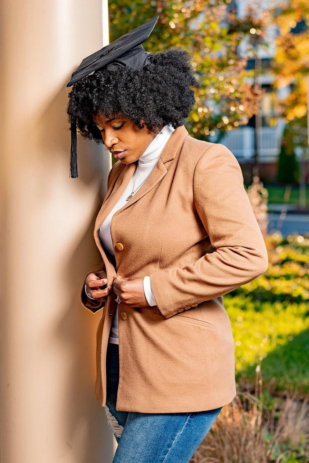 A woman with natural curly hair wearing a tan blazer, white turtleneck, and jeans, standing outdoors with a graduation cap hanging on her head, looking down and holding her blazer with both hands.