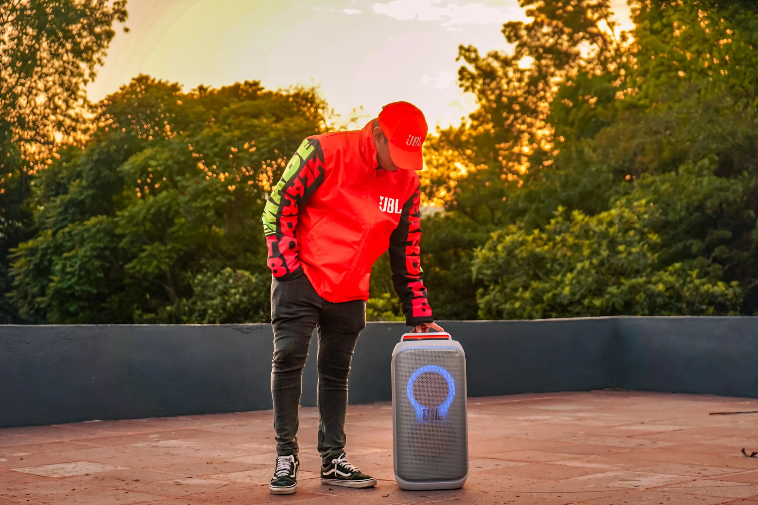 Un hombre con ropa deportiva roja con logotipo JBL y gorra JBL, agachado y tocando un altavoz portátil JBL en un ambiente al aire libre con árboles y un atardecer