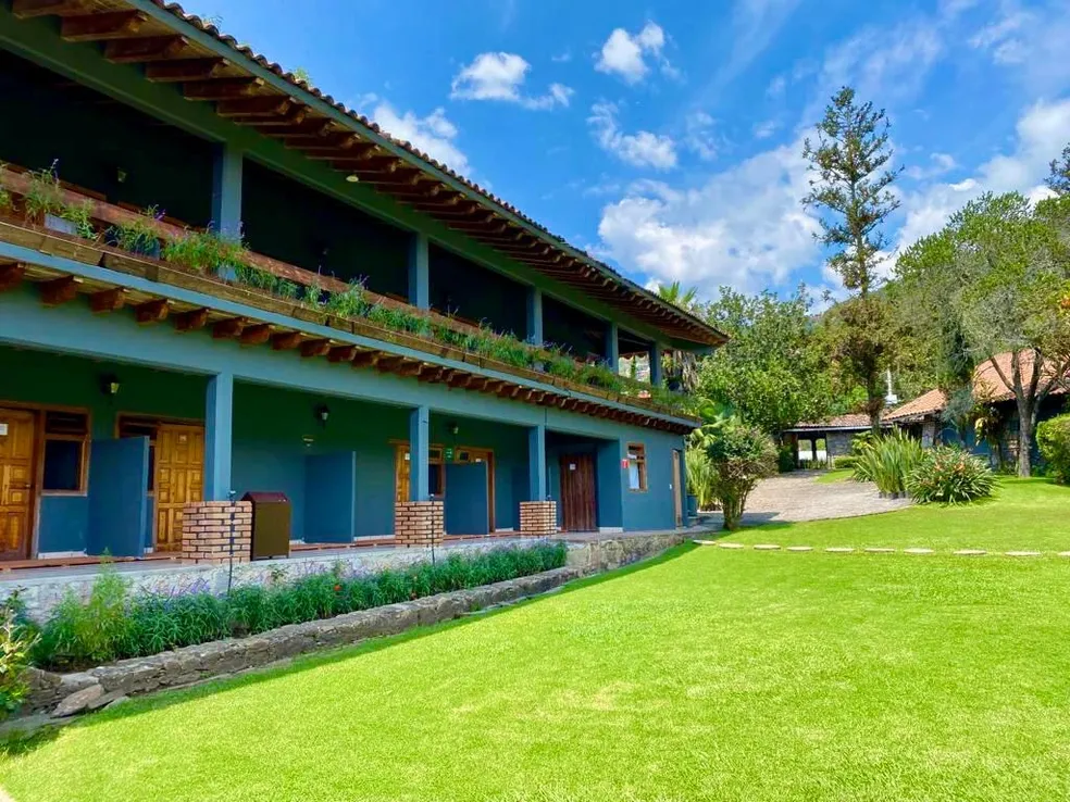 Edificio azul de dos pisos con balcón lleno de plantas, rodeado de césped verde y árboles, bajo un cielo azul con nubes.