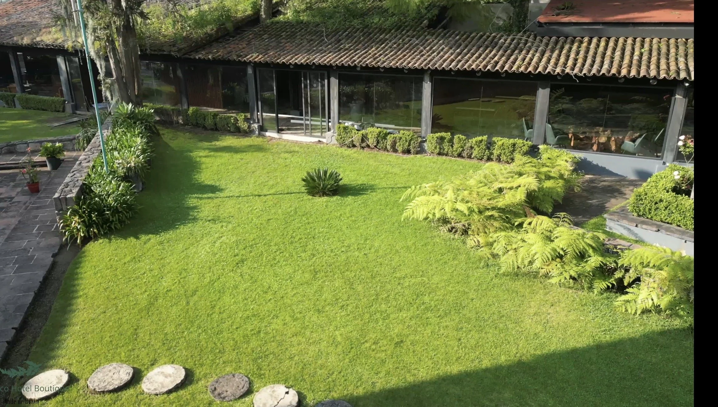 Vista de un jardín con césped bien cuidado, plantas en macetas y arbustos, y un edificio con ventanas grandes y un techo de tejas. El área parece ser parte de un restaurante o casa en un entorno natural.