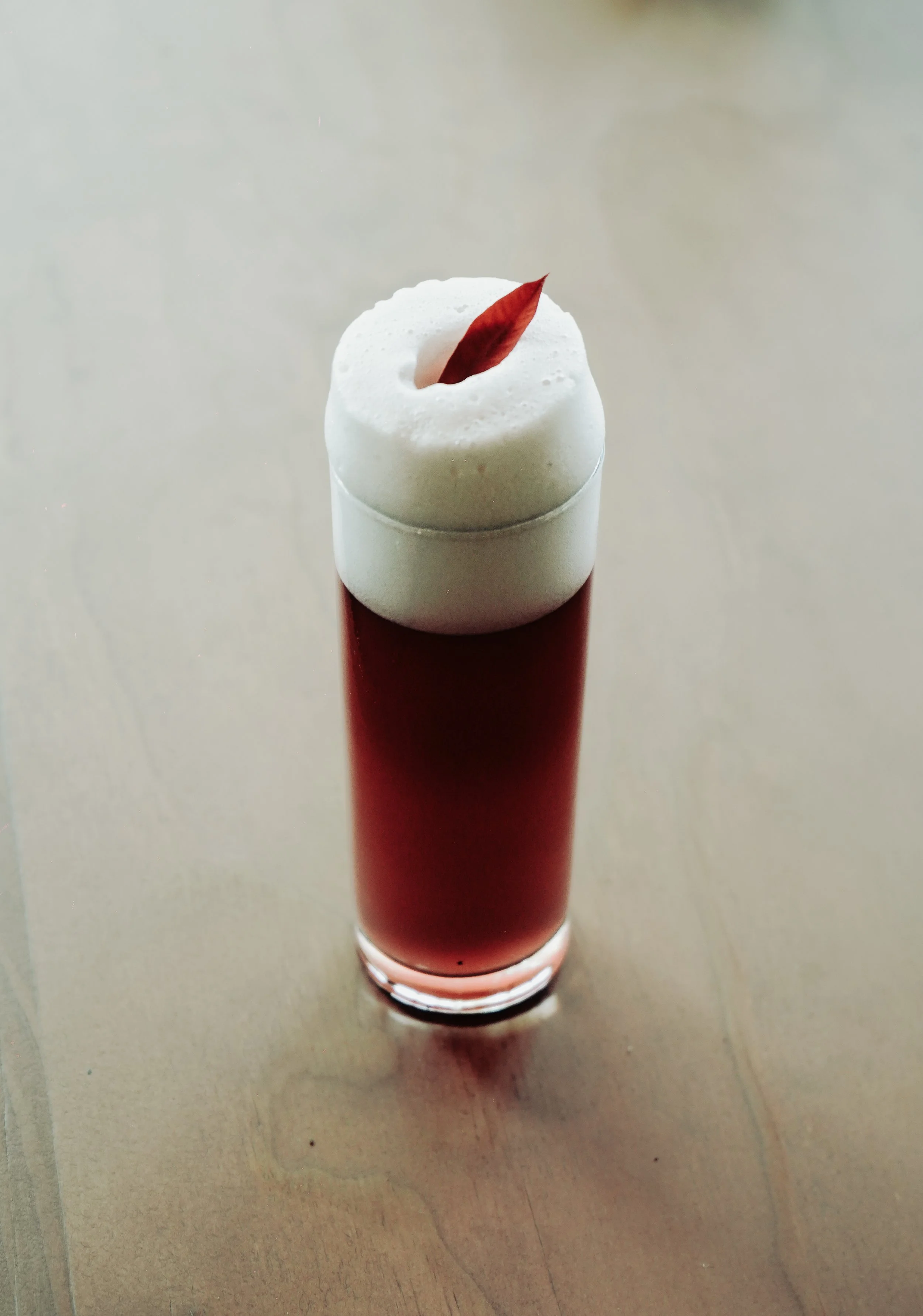 Vaso alto con bebida rojiza, cubierta con espuma blanca y decorada con una hoja roja.