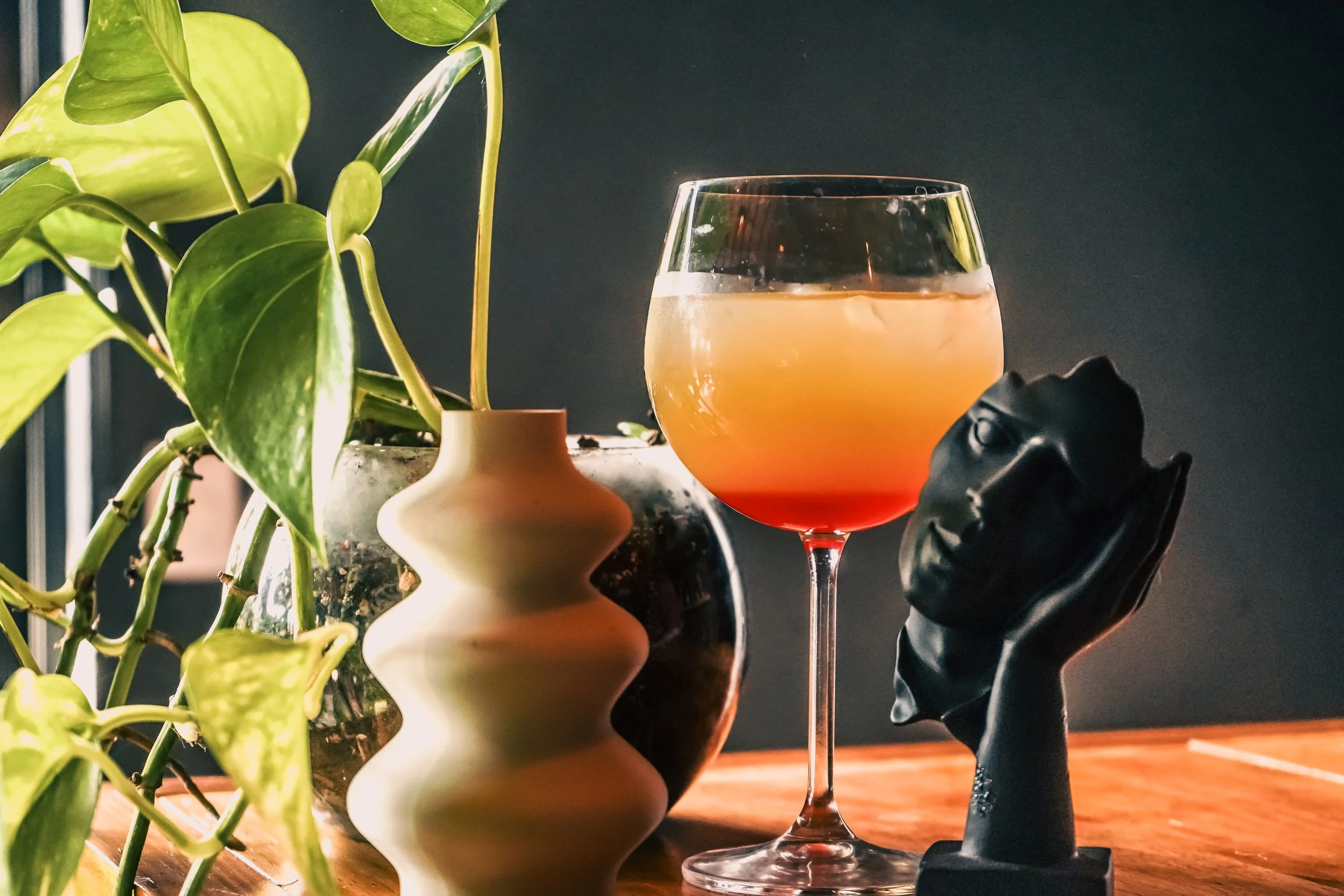 Una planta con hojas verdes en un jarrón de cerámica, una copa de vino rosado con un degradado de colores, y una escultura negra de una cabeza humana con una mano en la cara, en una superficie de madera con fondo oscuro.