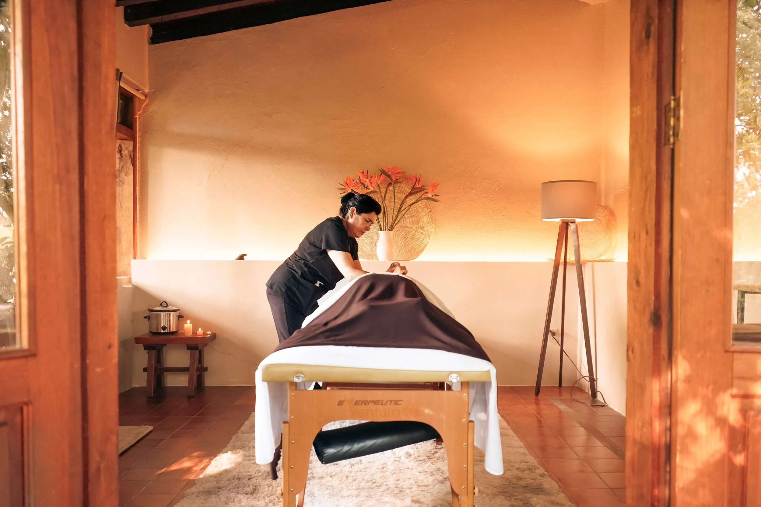 Una mujer recibe un masaje en una sala de spa con decoración cálida y luces suaves, en un ambiente relajante con una lámpara de pie y plantas decorativas.