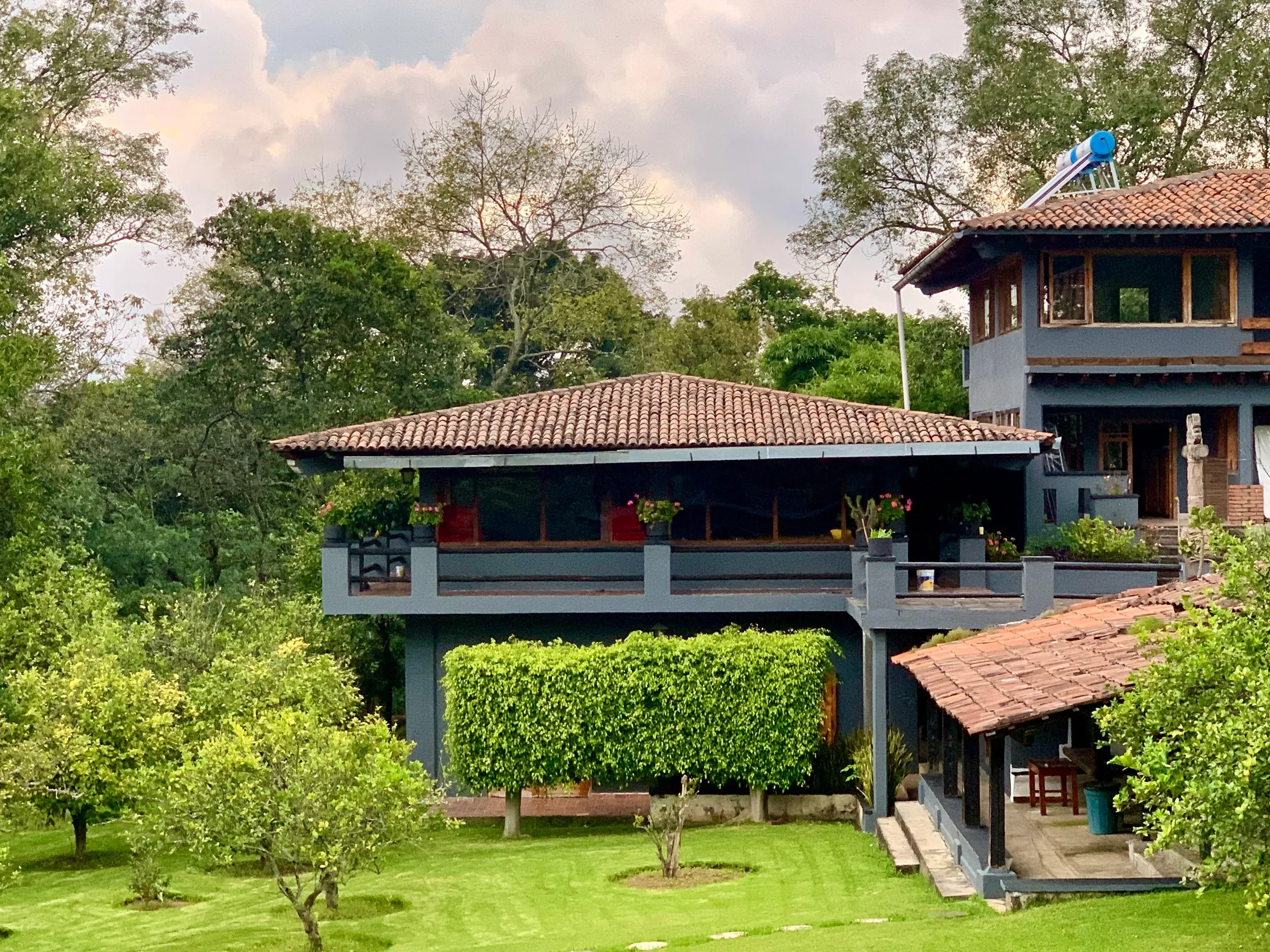 Casa de dos niveles con paredes azules, techo de tejas y balcón con plantas, rodeada de árboles y césped verde.