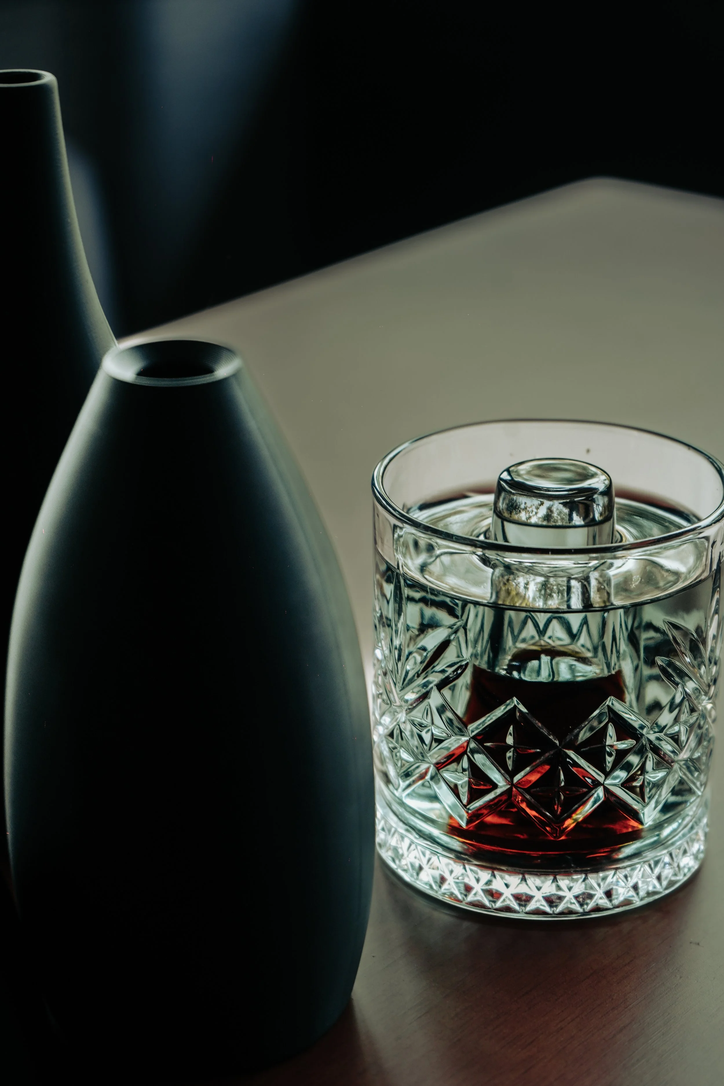 Vaso de cristal con whisky y hielo junto a dos jarrones negros en una mesa de madera.
