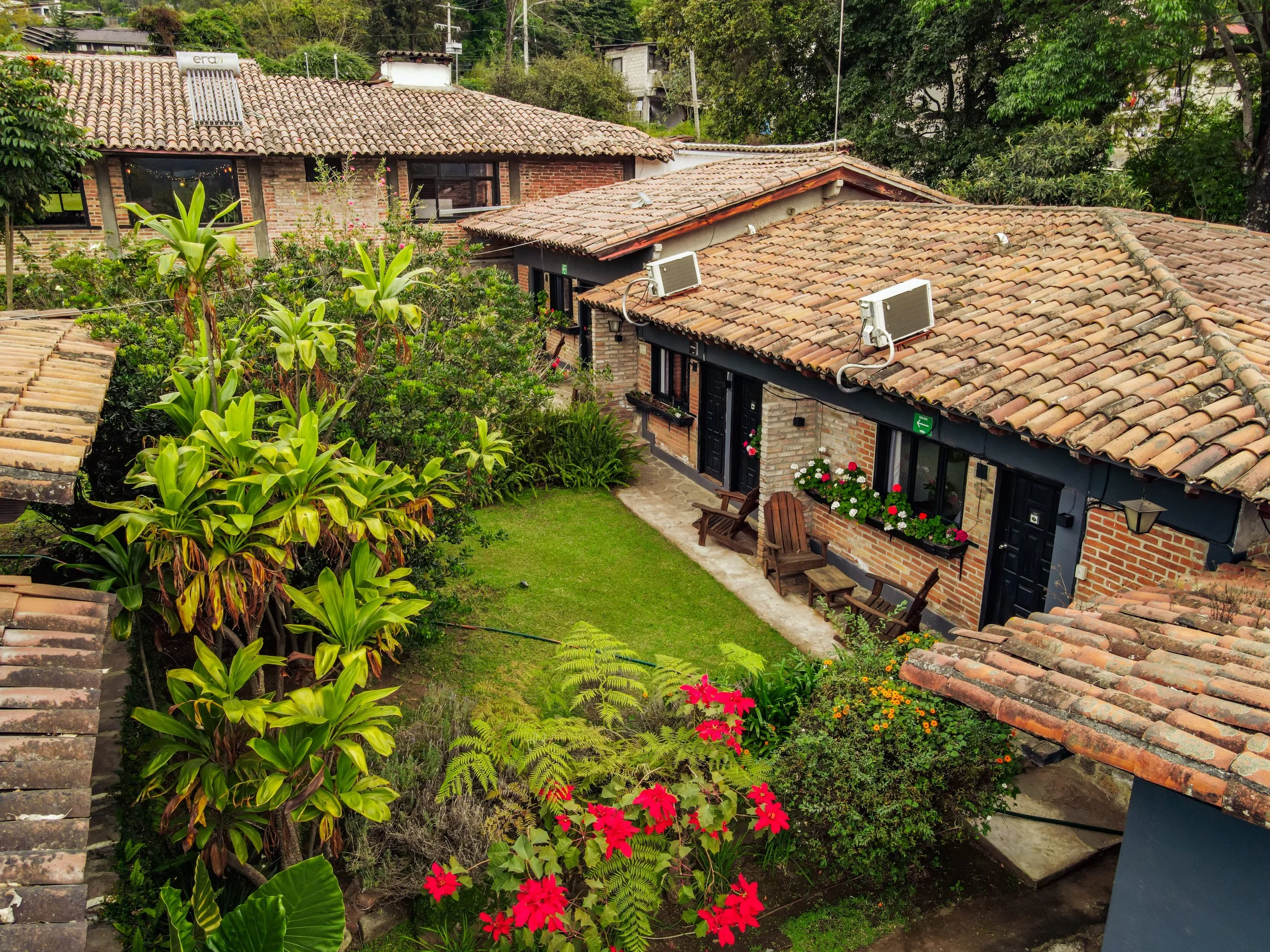 Cabañas con paredes de ladrillo, tejados de tejas, y aire acondicionado en el techo, rodeadas de jardín con flores y plantas verdes.