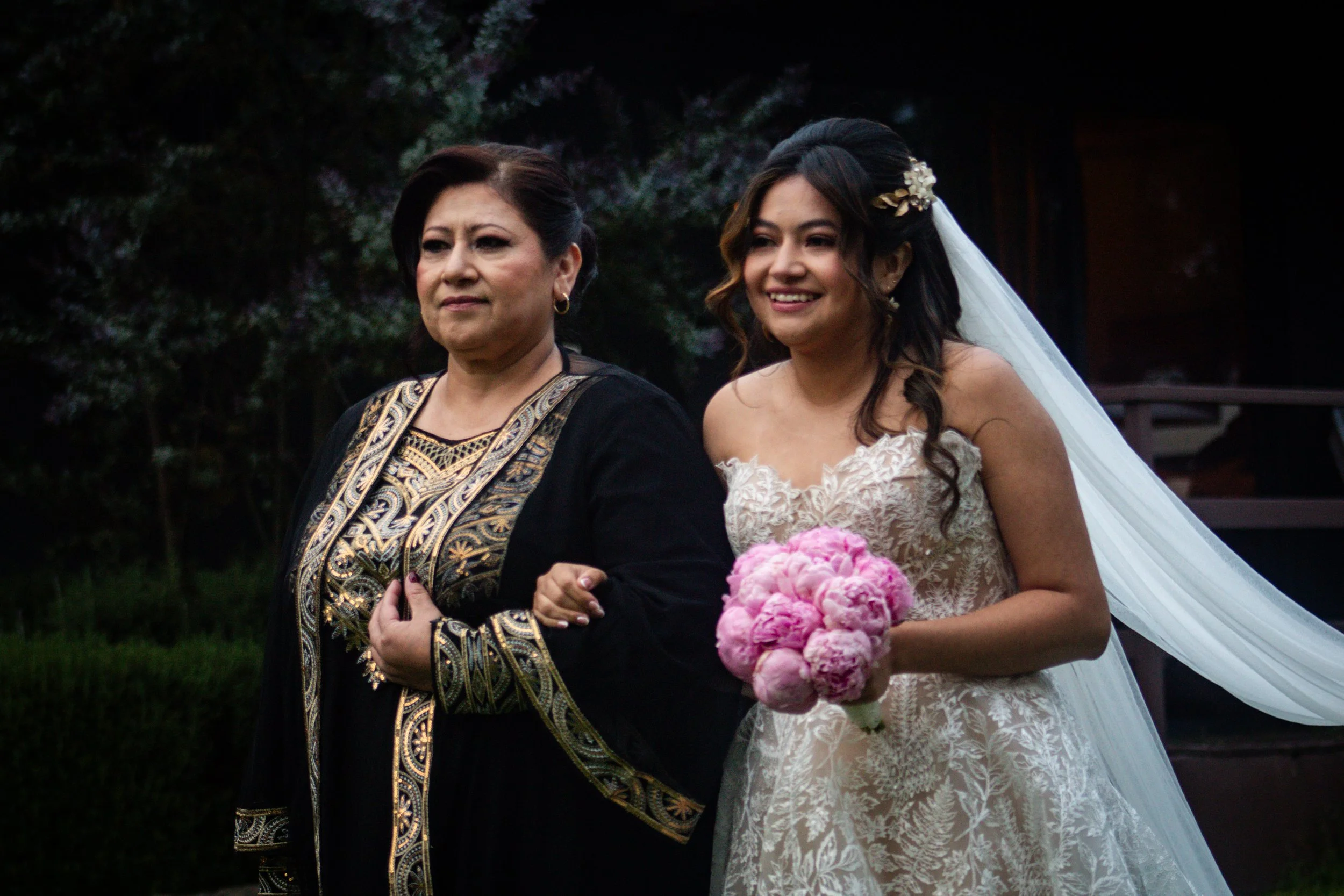 Mujer en vestido de novia sosteniendo ramo de flores rosas, acompañando a otra mujer vestida con ropa tradicional en un ambiente al aire libre.