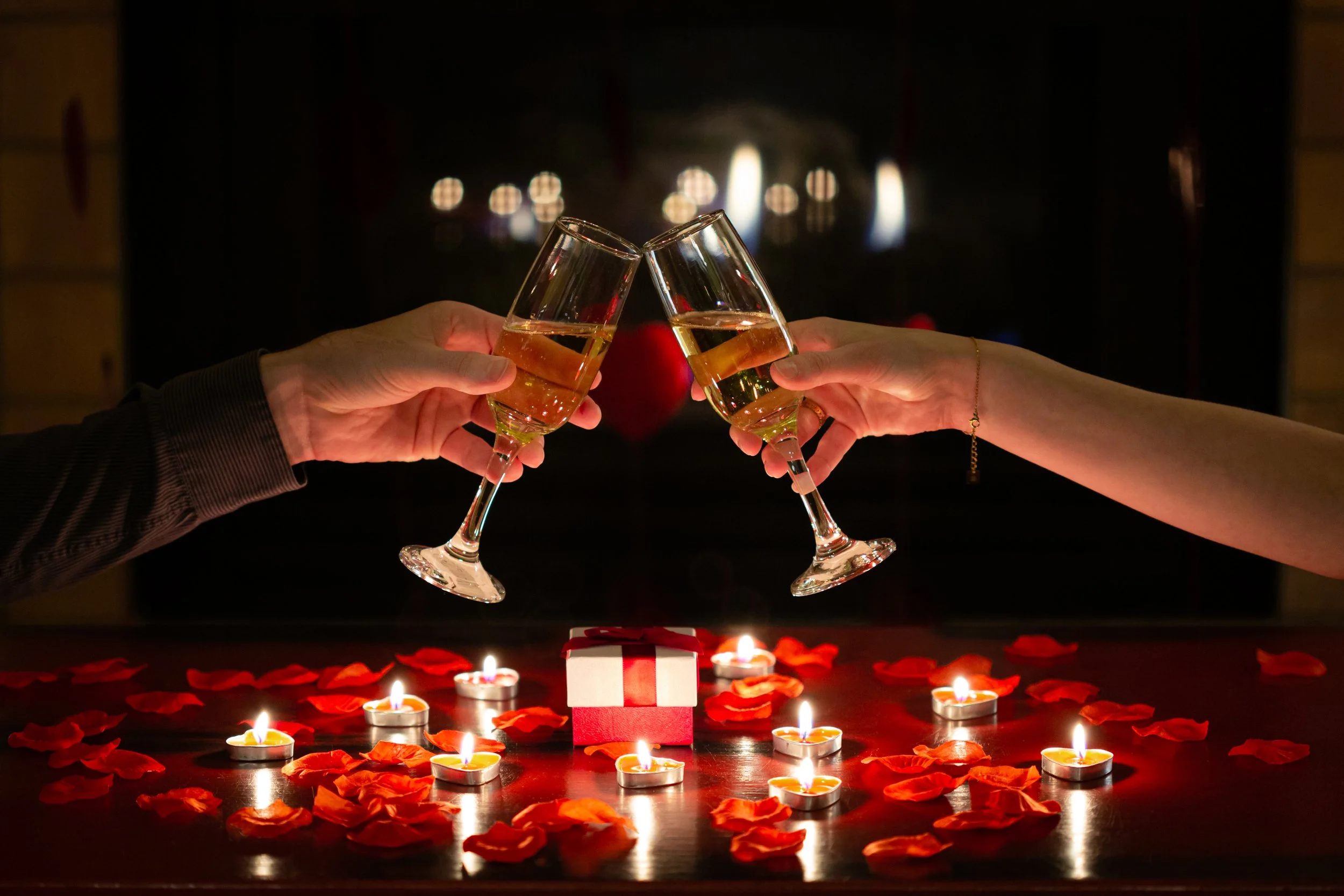 Pareja brindando con copas de champán en una cena romántica con velas y pétalos en la mesa.