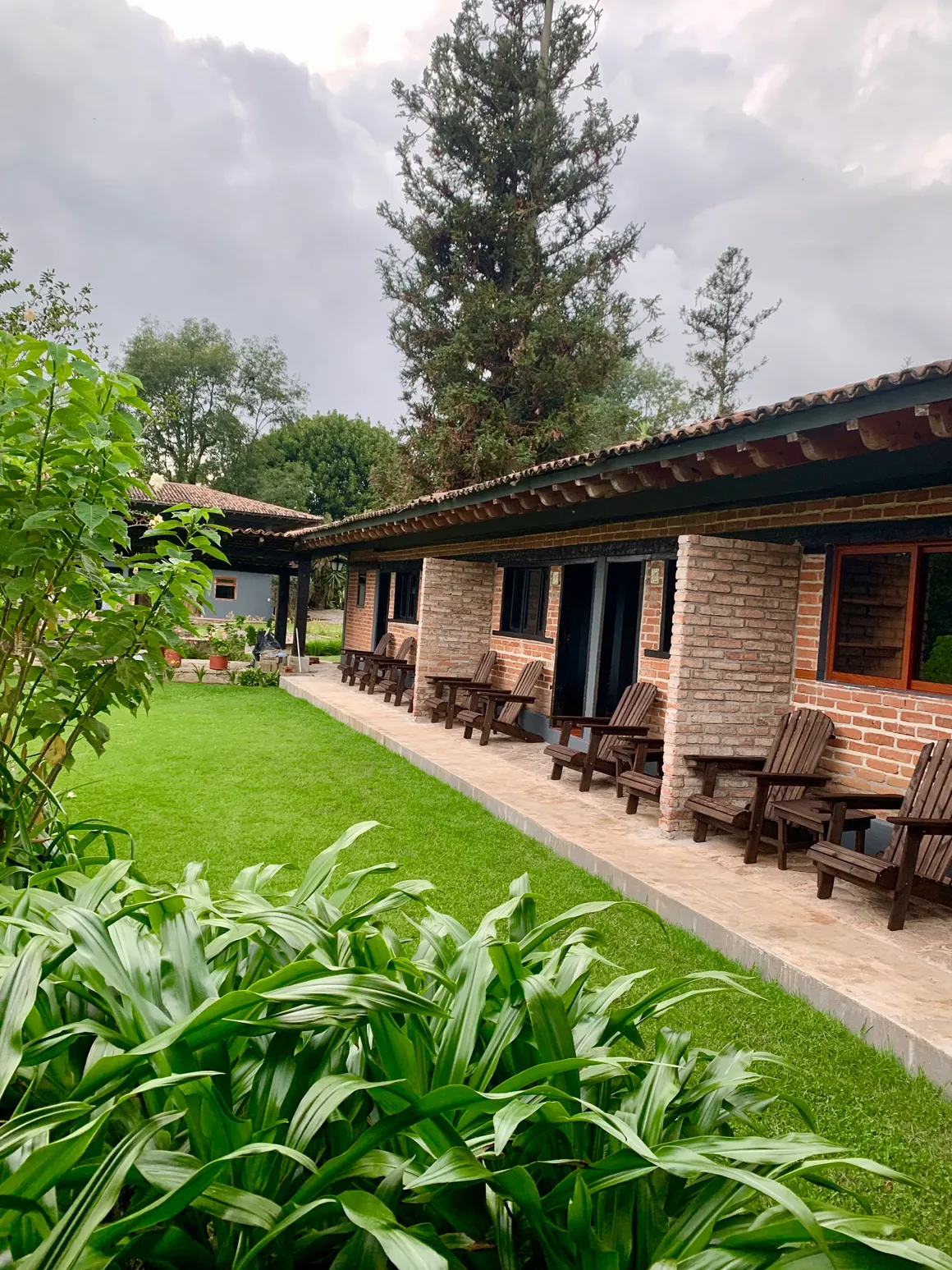 Vista de un edificio de una planta con muro de ladrillo y ventanas grandes, rodeado de vegetación, con muchas sillas de madera en el porche y un césped verde.