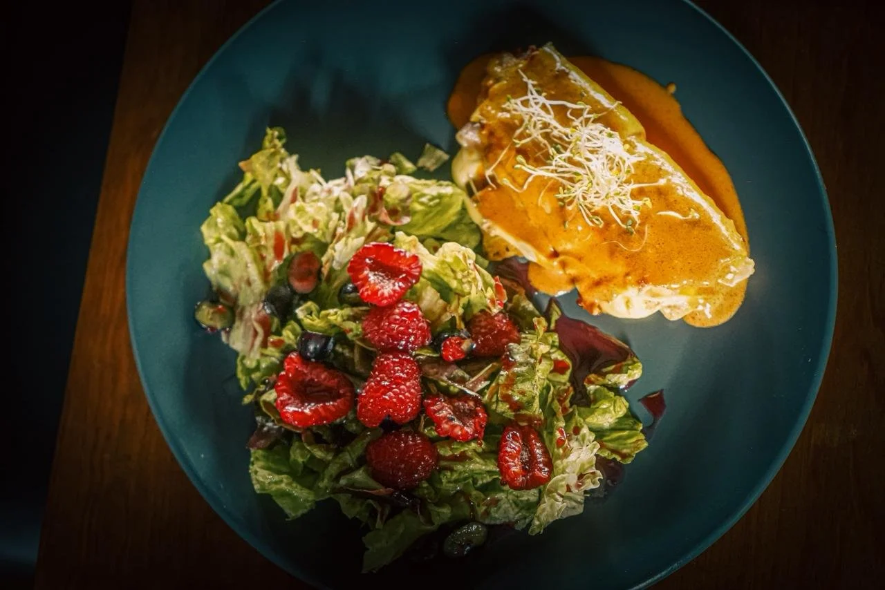 En un plato azul hay una ensalada mixta con frutas y un filete de pescado cubierto con salsa y adornado con brotes.