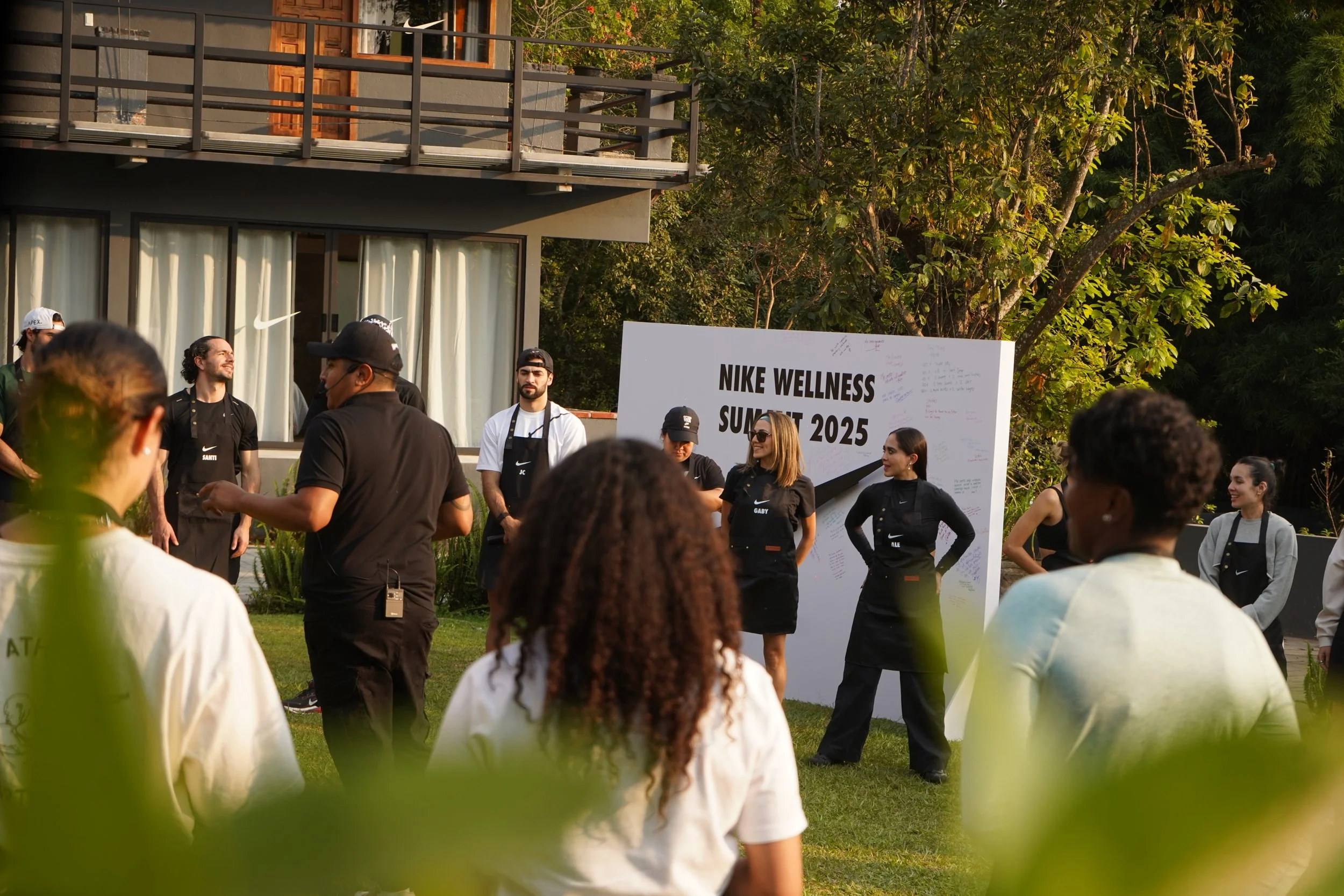 Grupo de personas participando en un evento de Nike Wellness Summit 2025 al aire libre, con un cartel grande que dice y con árboles y una casa moderna de fondo.