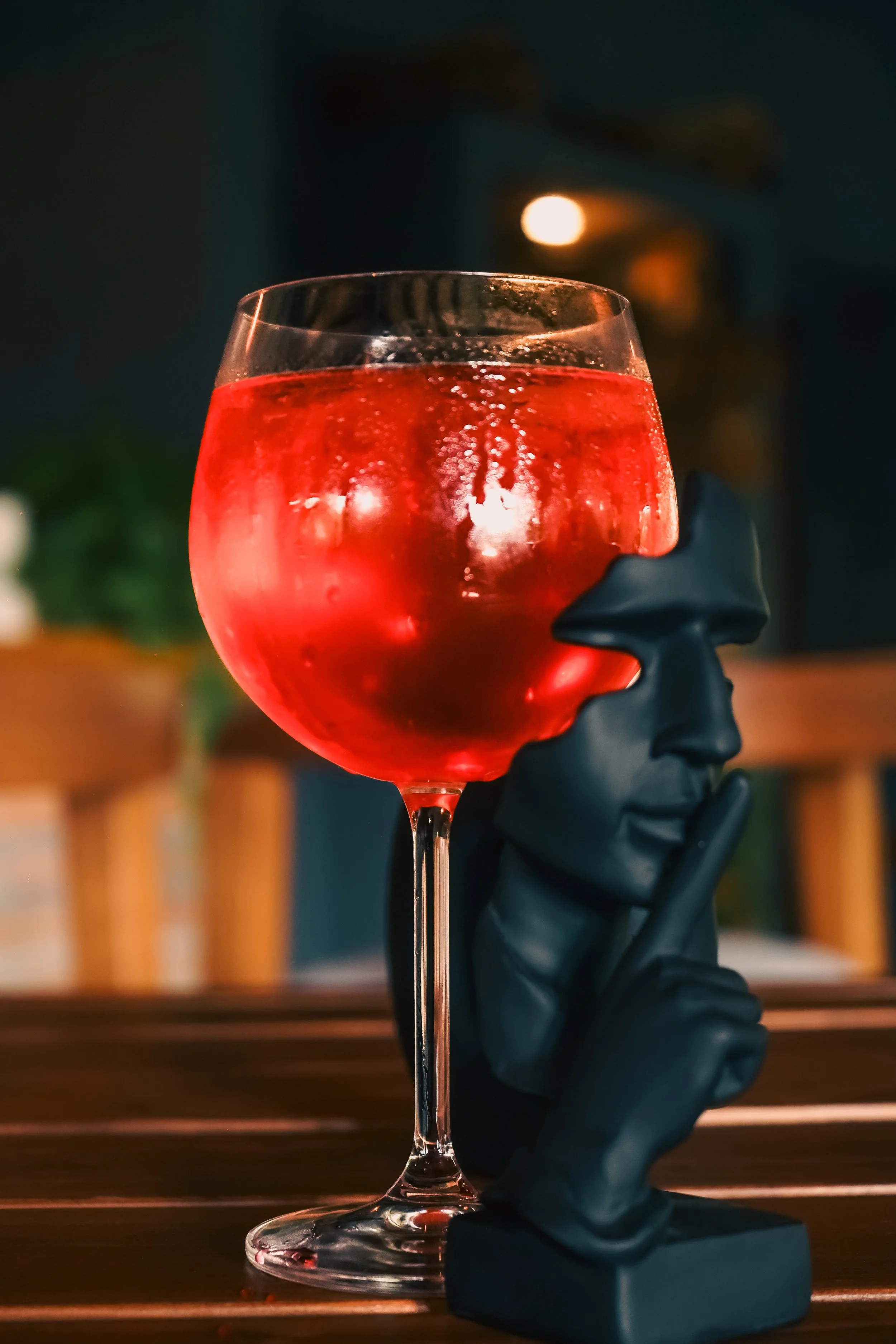 Copa de cóctel rojo sobre una mesa de madera, con una escultura negra de una cara humana con un dedo en los labios, fondo borroso con luces y plantas.