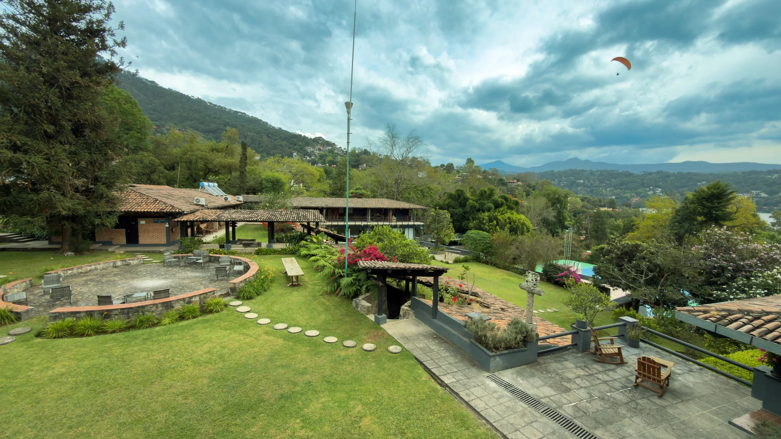 Jardín con césped, caminos de piedra, árboles y plantas, estructura de techos de tejas, paisaje montañoso y cielo nublado, con parapente en el aire.
