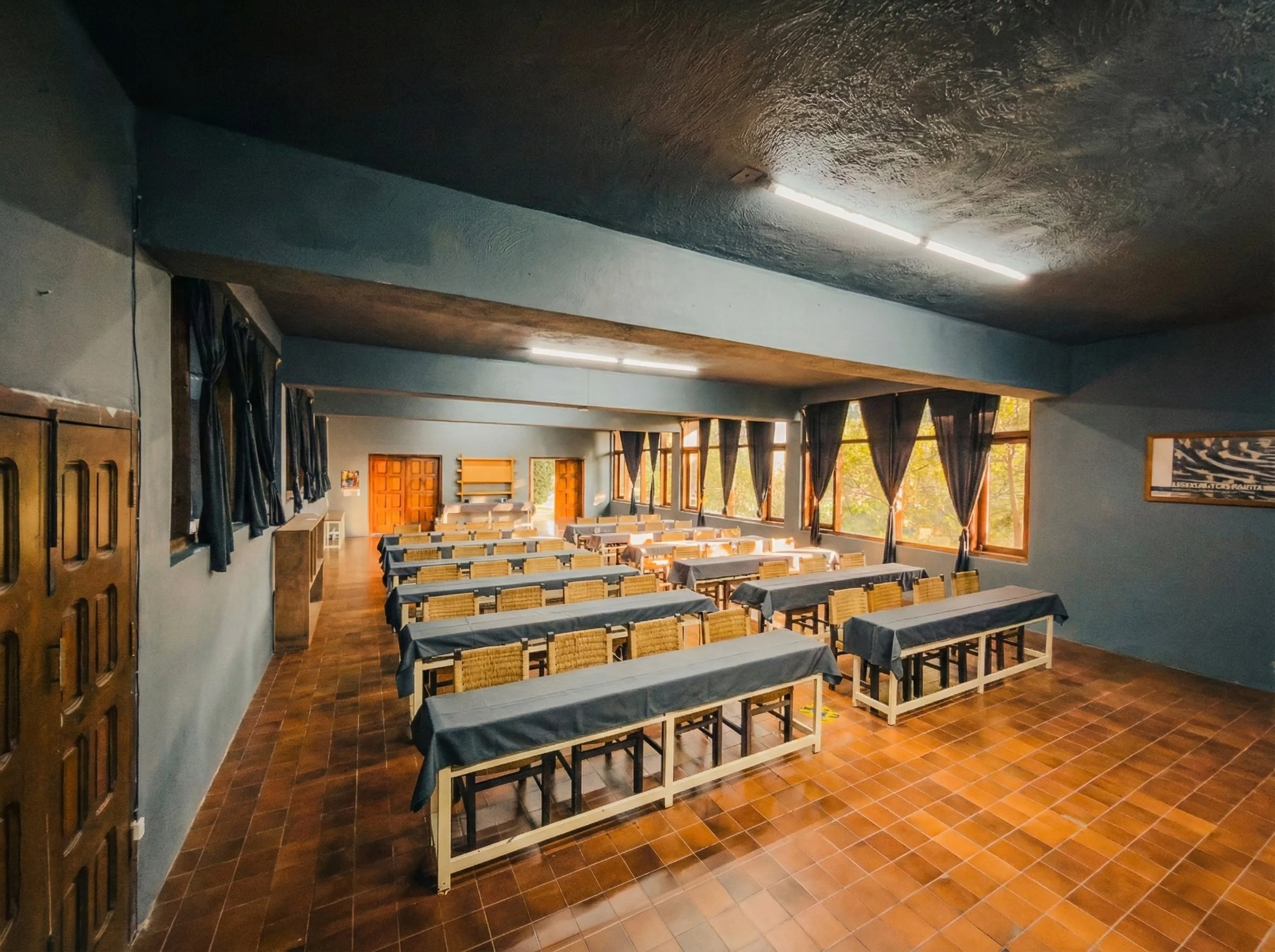 Habitación de hotel con cama grande, cabecera de madera, paredes grises, techo de madera expuesta, muebles de madera, sillón de cuero, piso de madera, luz cálida y una puerta que lleva a un baño.