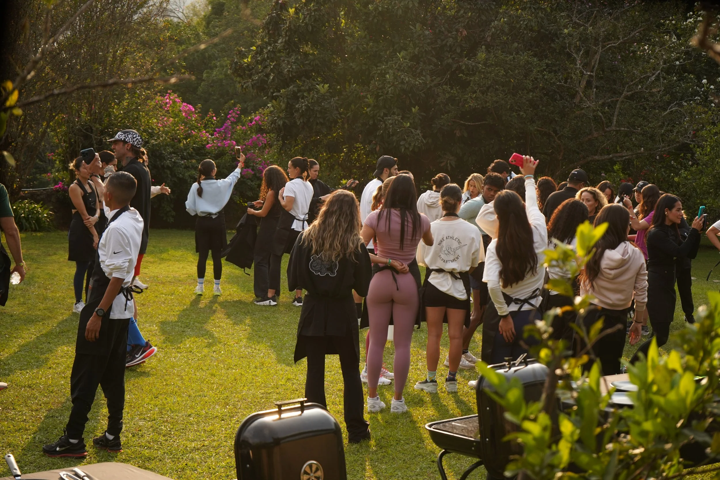 Varias personas en un jardín al atardecer, algunas tomando fotos y otras conversando, rodeados de árboles y flores coloridas.