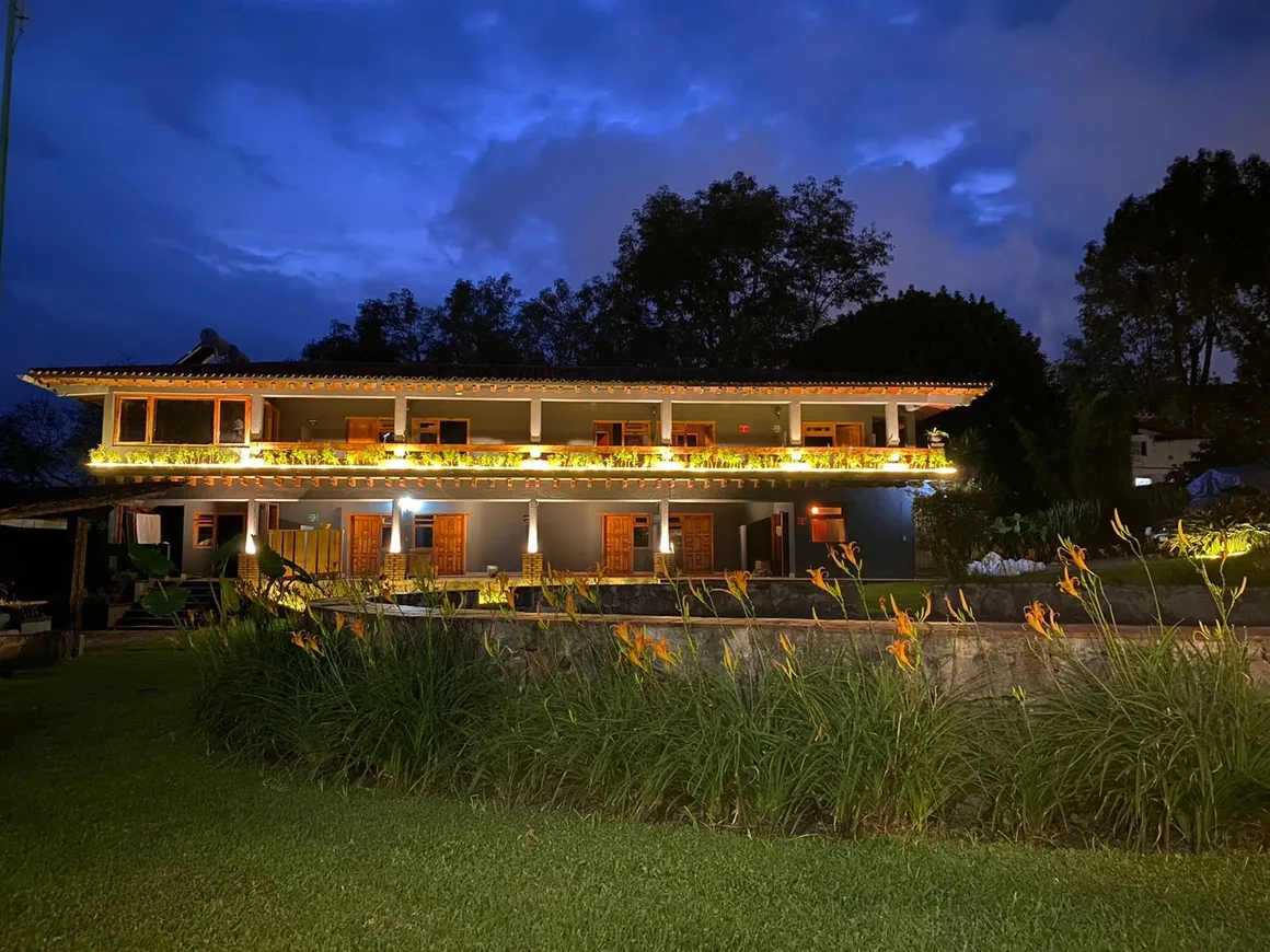 Edificio residencial de dos pisos iluminado por la noche, rodeado de jardín con flores y árboles.