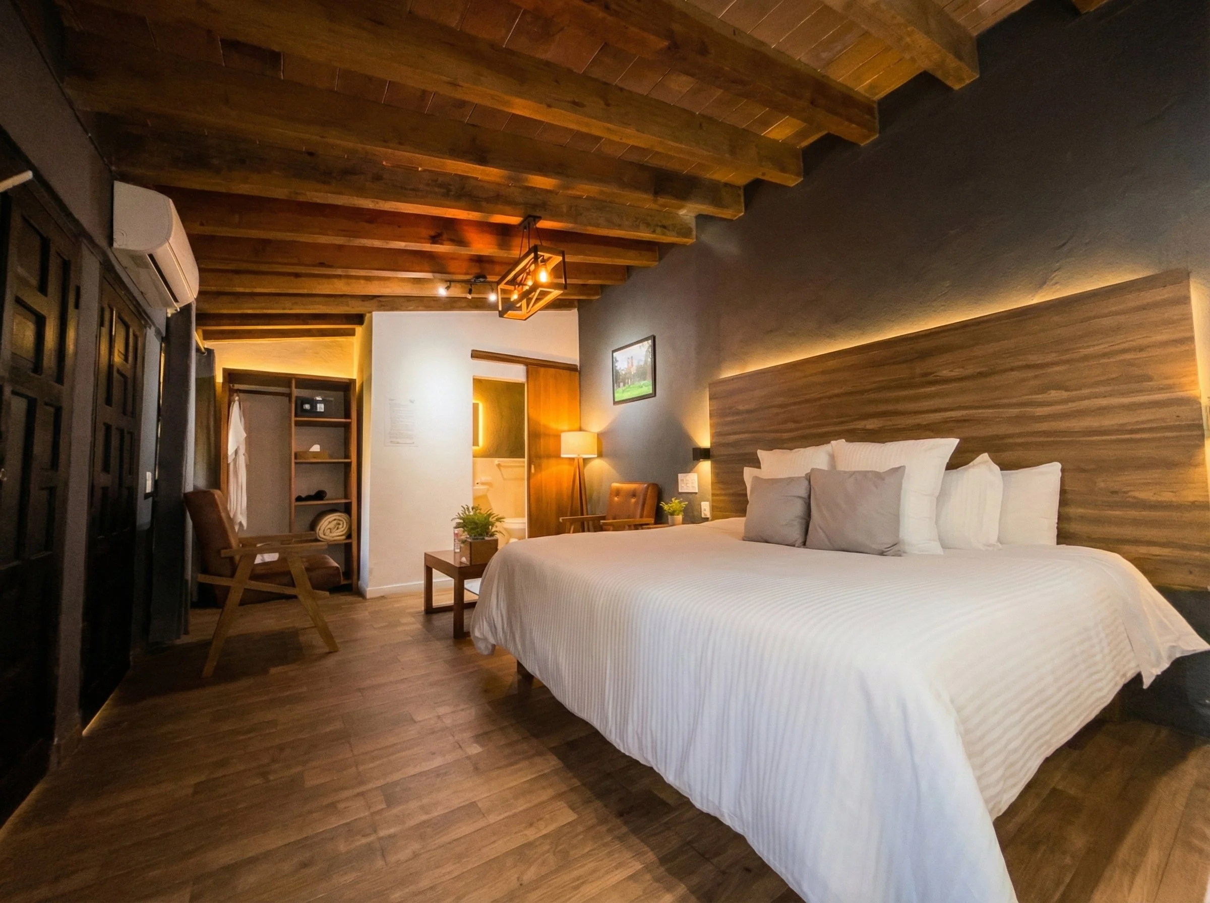 Habitación de hotel con cama grande, cabecera de madera, lámparas de lectura, sillón, mesa de noche con plantas, paredes de color gris y madera, techo con vigas de madera, interior cálido y moderno.