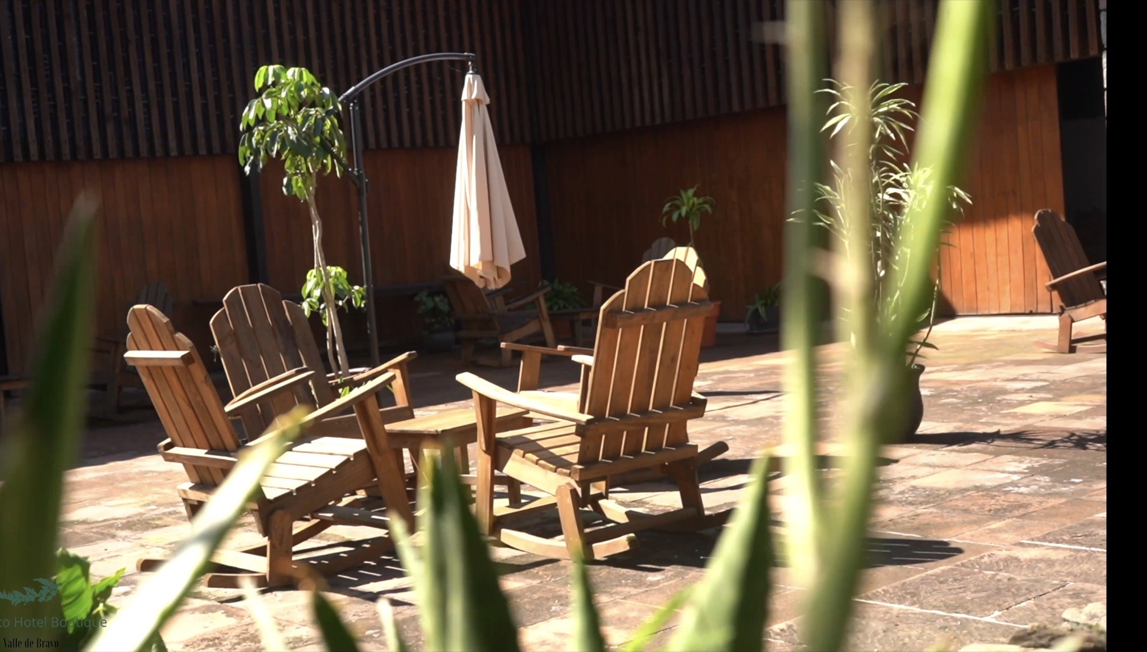 Área de descanso con sillas de madera, una sombrilla, plantas en macetas y un muro de madera en un espacio exterior.