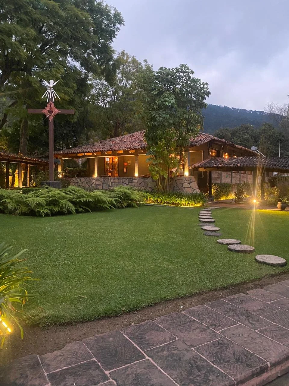 Edificio de dos plantas con ventanas largas, balcón en la segunda planta, rodeado de árboles y plantas, con área de descanso con sillas y mesa en el jardín.