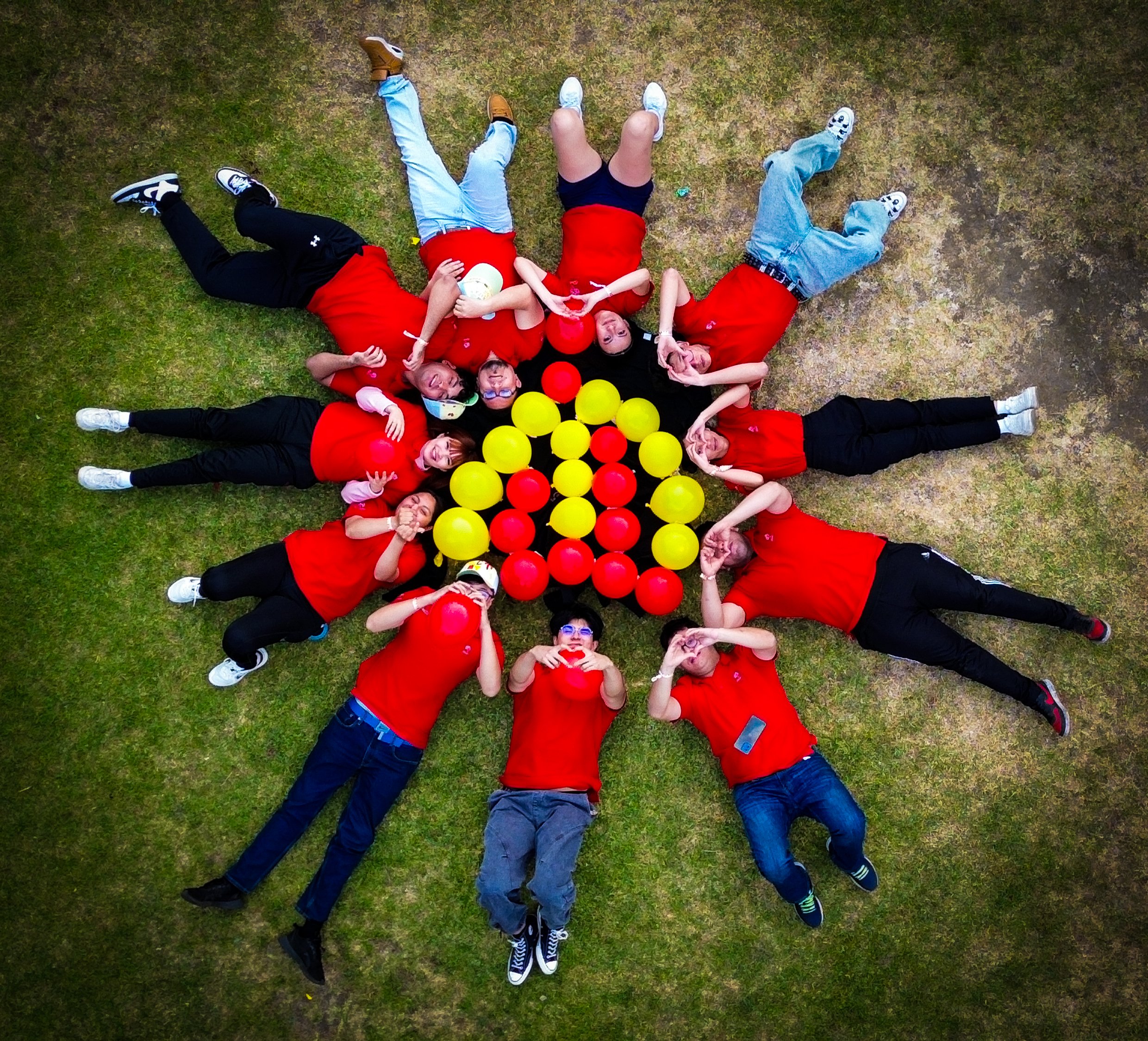 Grupo de personas en círculos en el césped, rodeando globos rojos y amarillos, algunas hacen gestos divertidos y todos llevan camisetas rojas.
