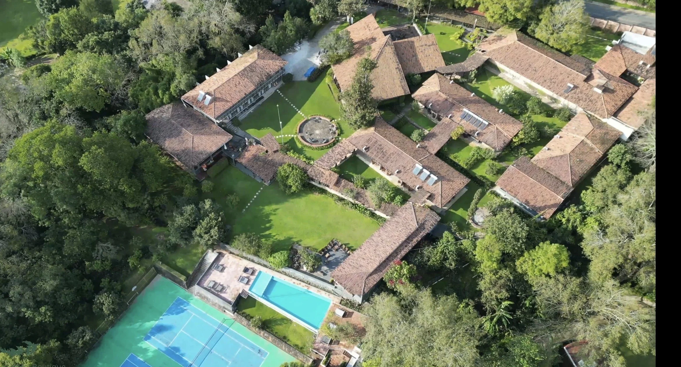 Vista aérea de un complejo de construcciones con techos de teja, áreas verdes, una piscina, y una cancha de tenis, rodeado de árboles.