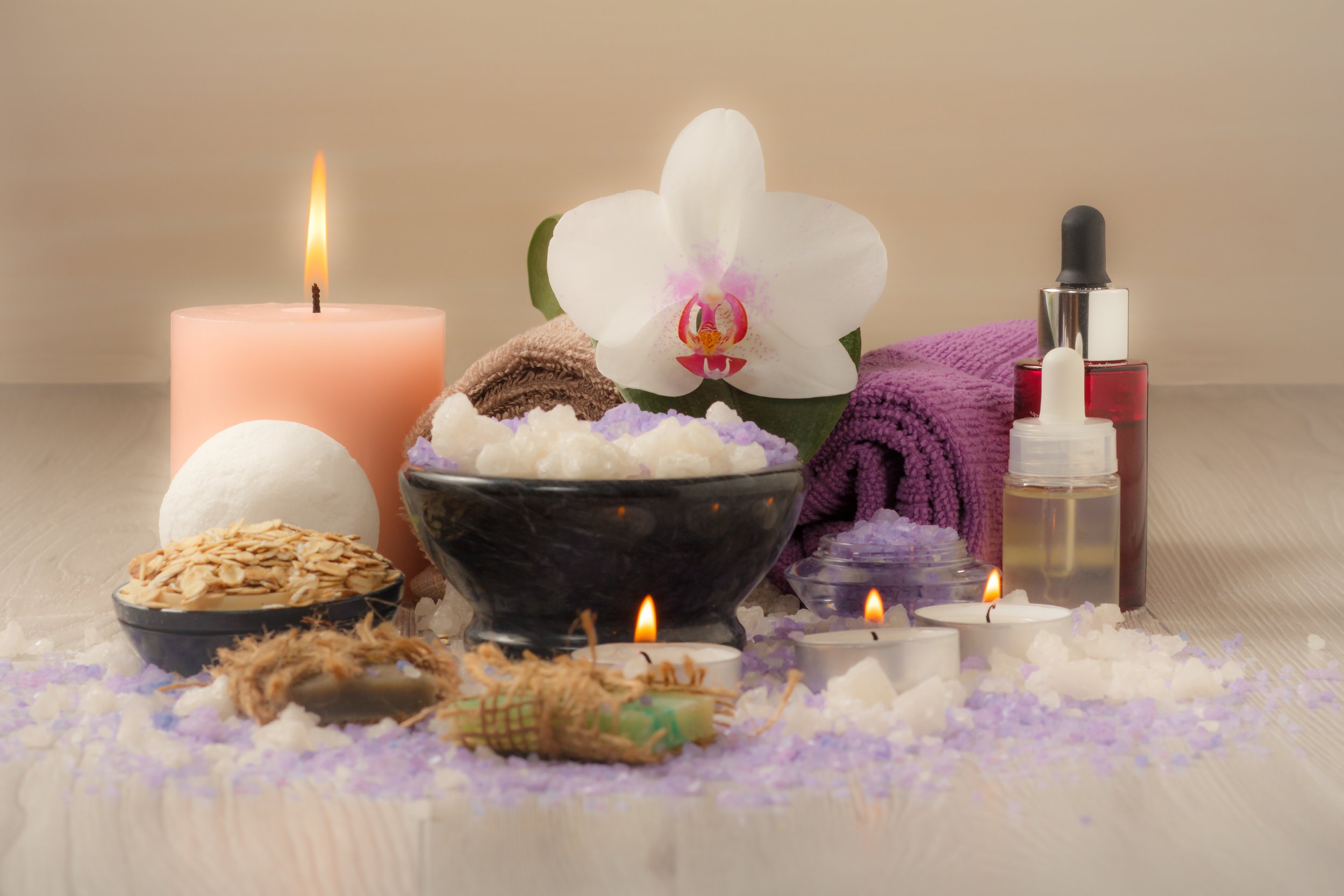 Configuración de spa con vela, flor de orquídea, toallas moradas, aceites, margaritas, y piedras decorativas.