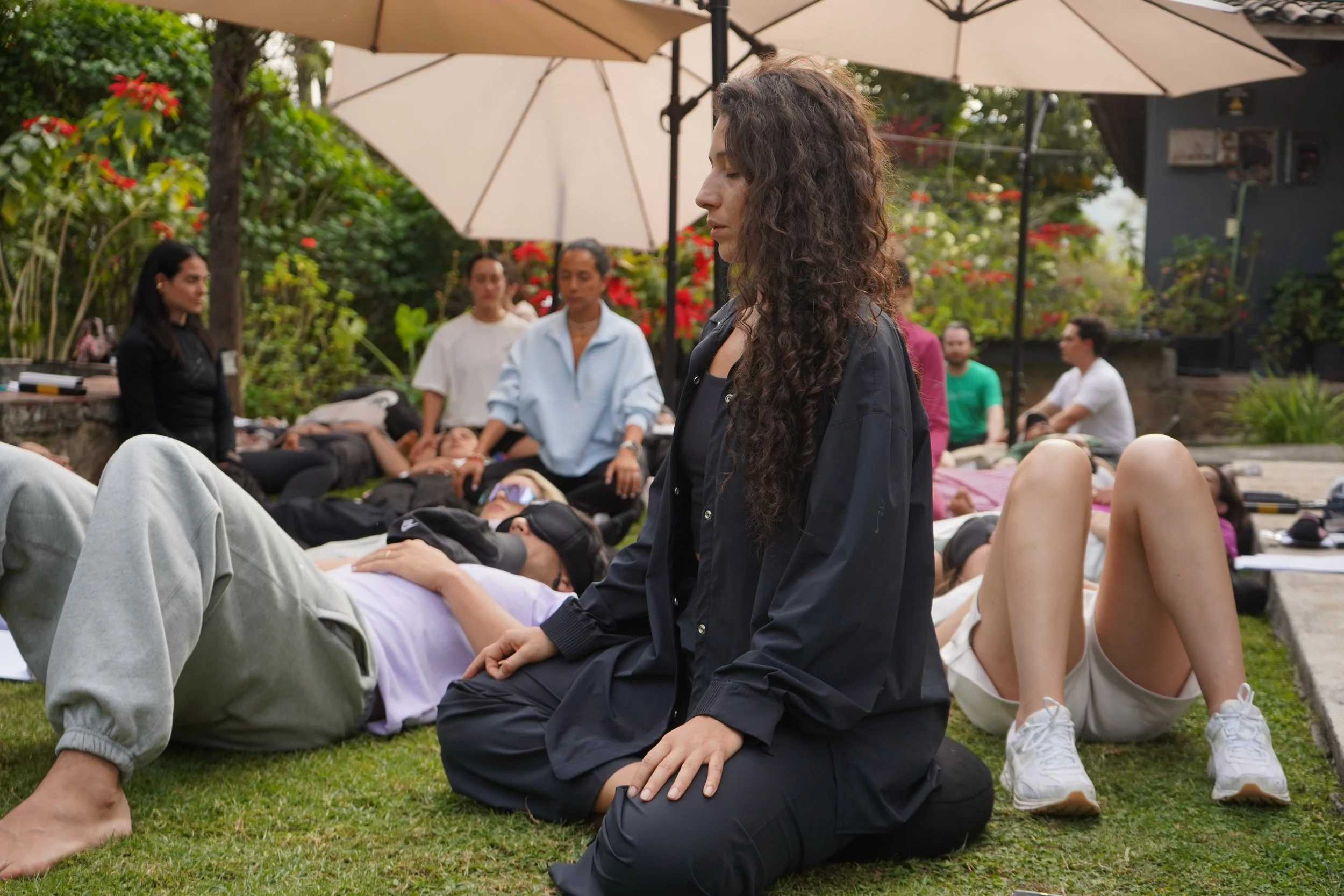 Grupo de personas en una ceremonia de meditación al aire libre, con varios descansando en el césped y un lugar rodeado de plantas y árboles.
