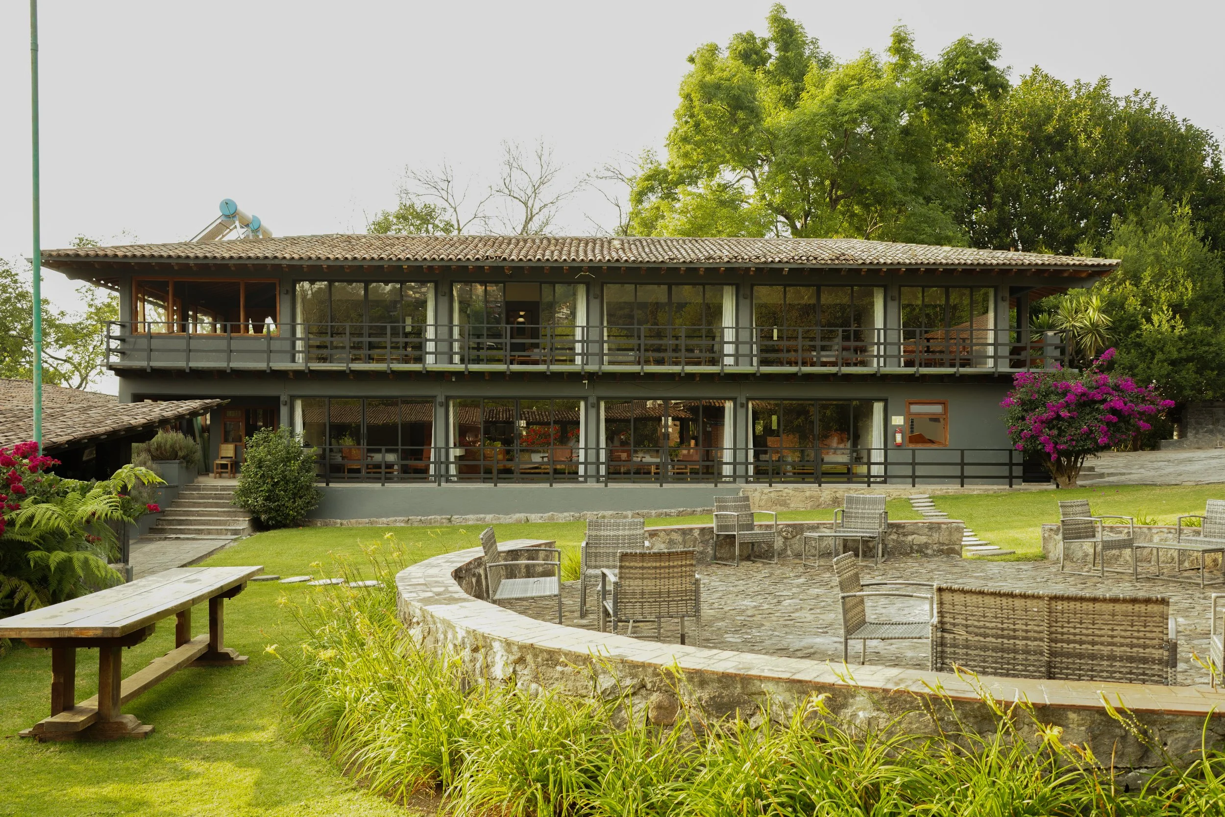 Edificio de dos plantas con ventanas largas, balcón en la segunda planta, rodeado de árboles y plantas, con área de descanso con sillas y mesa en el jardín.