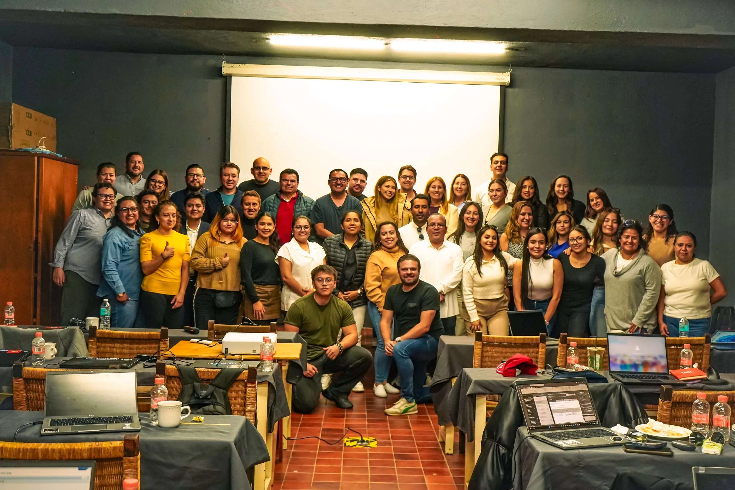 Grupo grande de personas sonriendo y posando para una foto en una sala de reuniones o conferencia, con mesas, laptops y botellas de agua en primer plano.