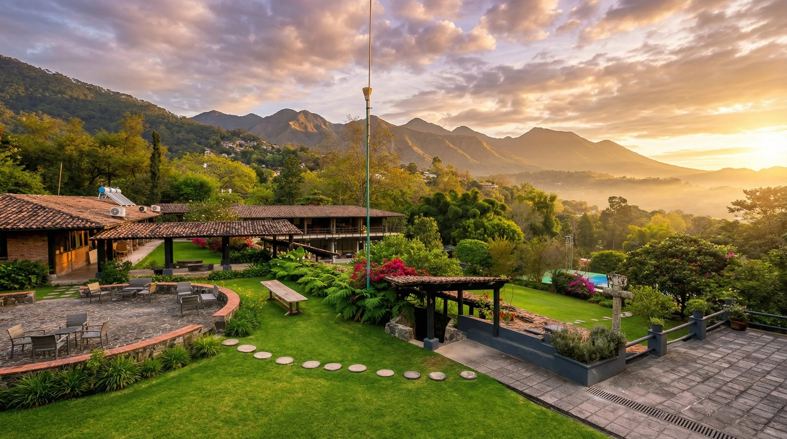Jardín con césped, caminos de piedra, árboles y plantas, estructura de techos de tejas, paisaje montañoso y cielo nublado, con parapente en el aire.