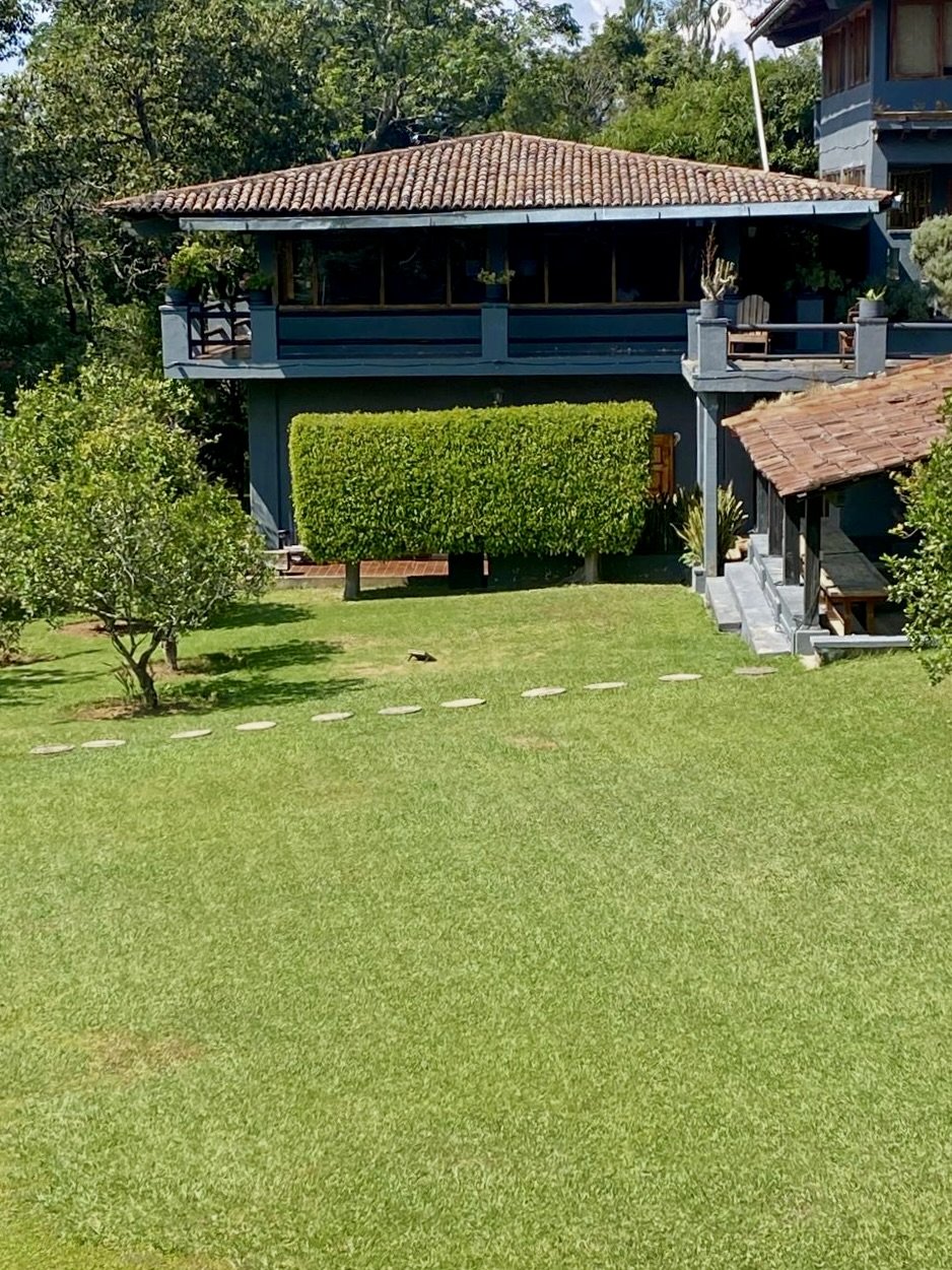 Jardín con césped, caminos de piedra, árboles y plantas, estructura de techos de tejas, paisaje montañoso y cielo nublado, con parapente en el aire.